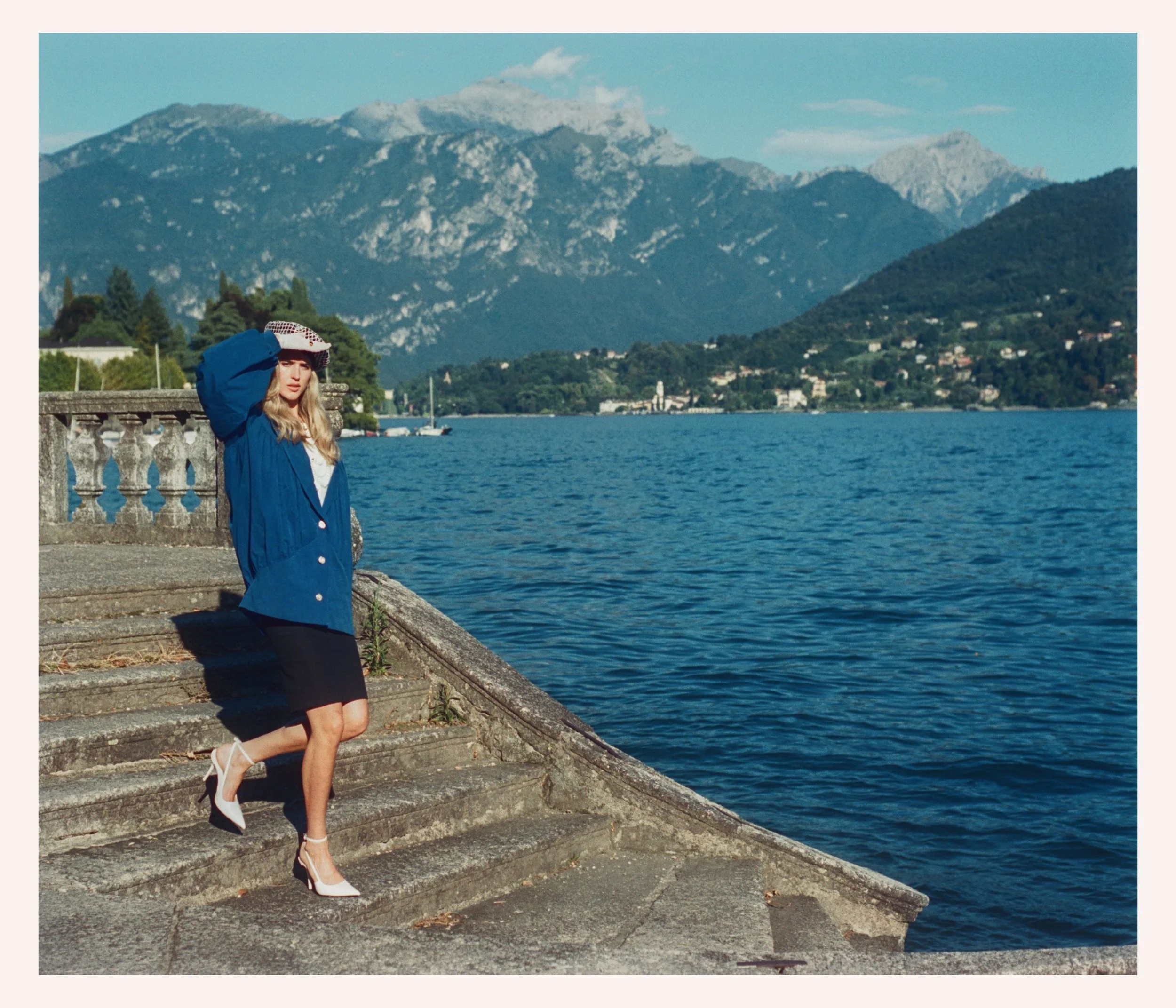 A woman standing on stone steps by a lake with mountains in the background. She is wearing a large blue jacket, a black skirt, white heels, and a wide-brimmed hat, and is shielding her eyes from the sun with her hand.