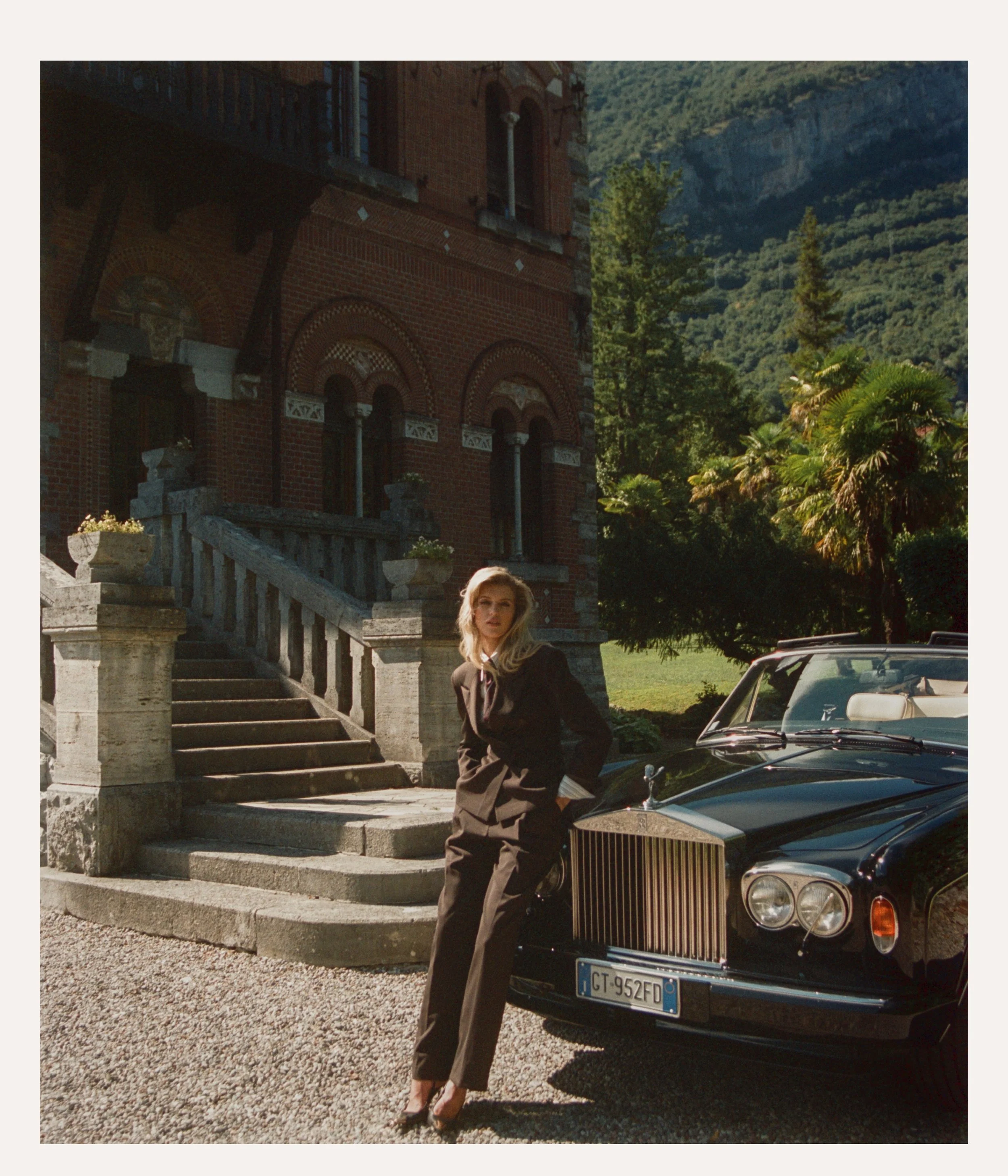 A woman in a black suit posing next to a classic black Rolls Royce car parked in front of a historic brick building with stairs and arches, surrounded by lush greenery and tall trees.