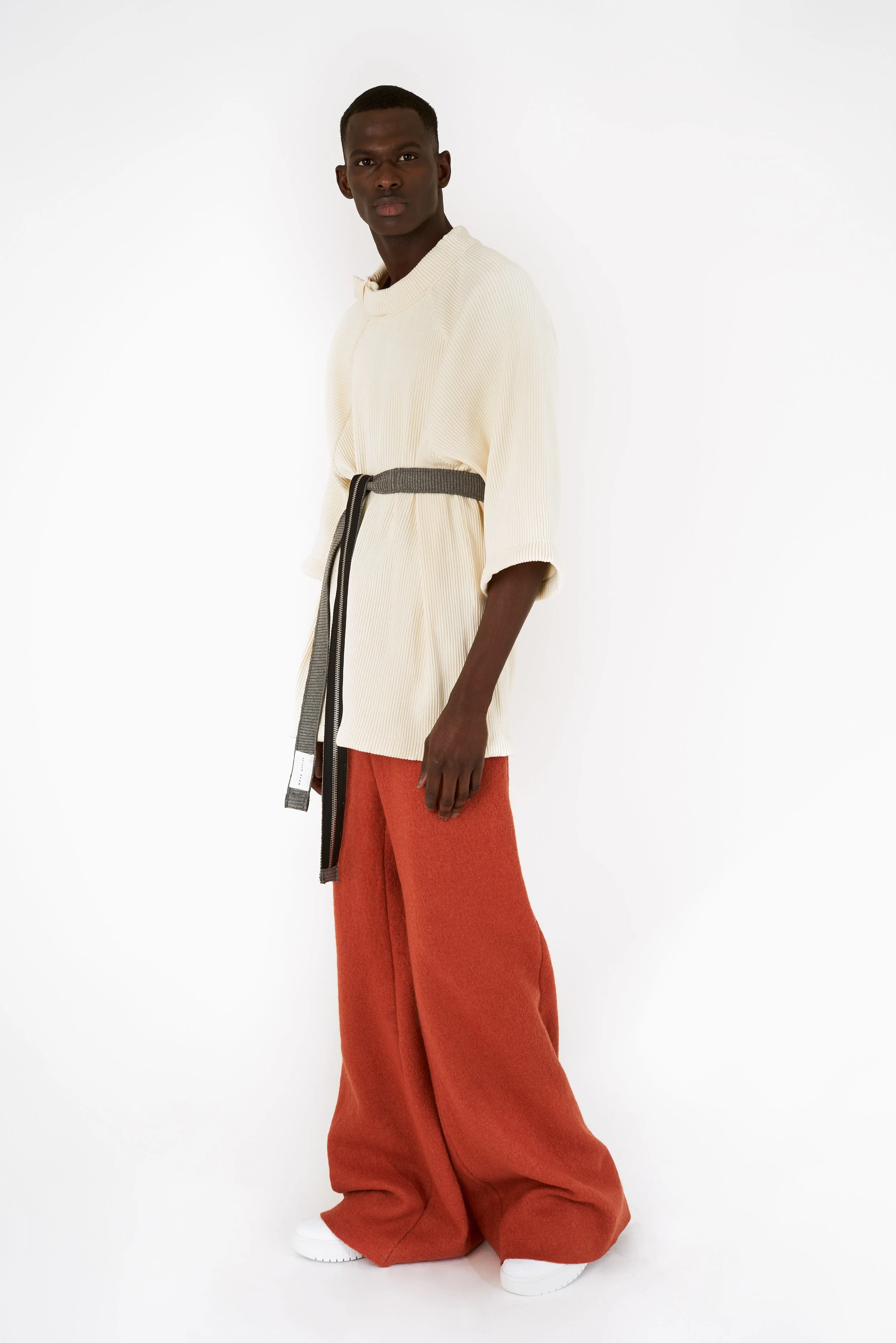 A tall person with dark skin and short hair wearing a cream-colored oversized top with 3/4 sleeves, wide orange pants, a gray belt, and white sneakers, standing against a white background.