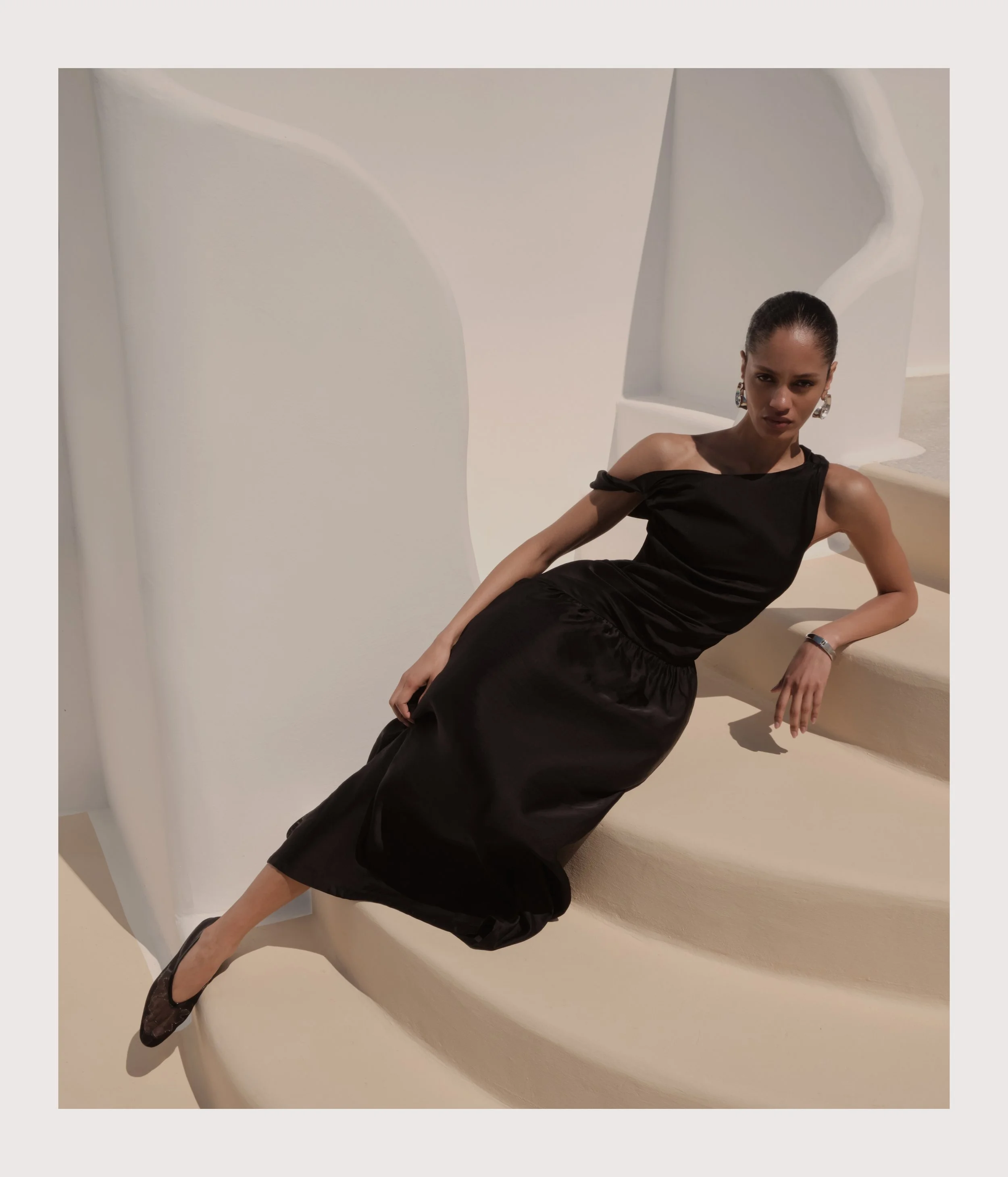 A woman in a black dress sitting on a curved, cream-colored staircase with white, abstract architectural background.