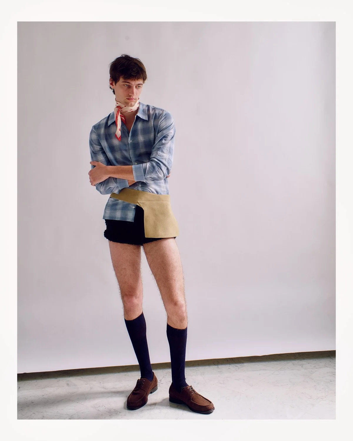 A young man standing against a plain white background wearing a plaid shirt, shorts, knee-high socks, and dress shoes, with a colorful pouch hanging from his waist and a scarf around his neck.