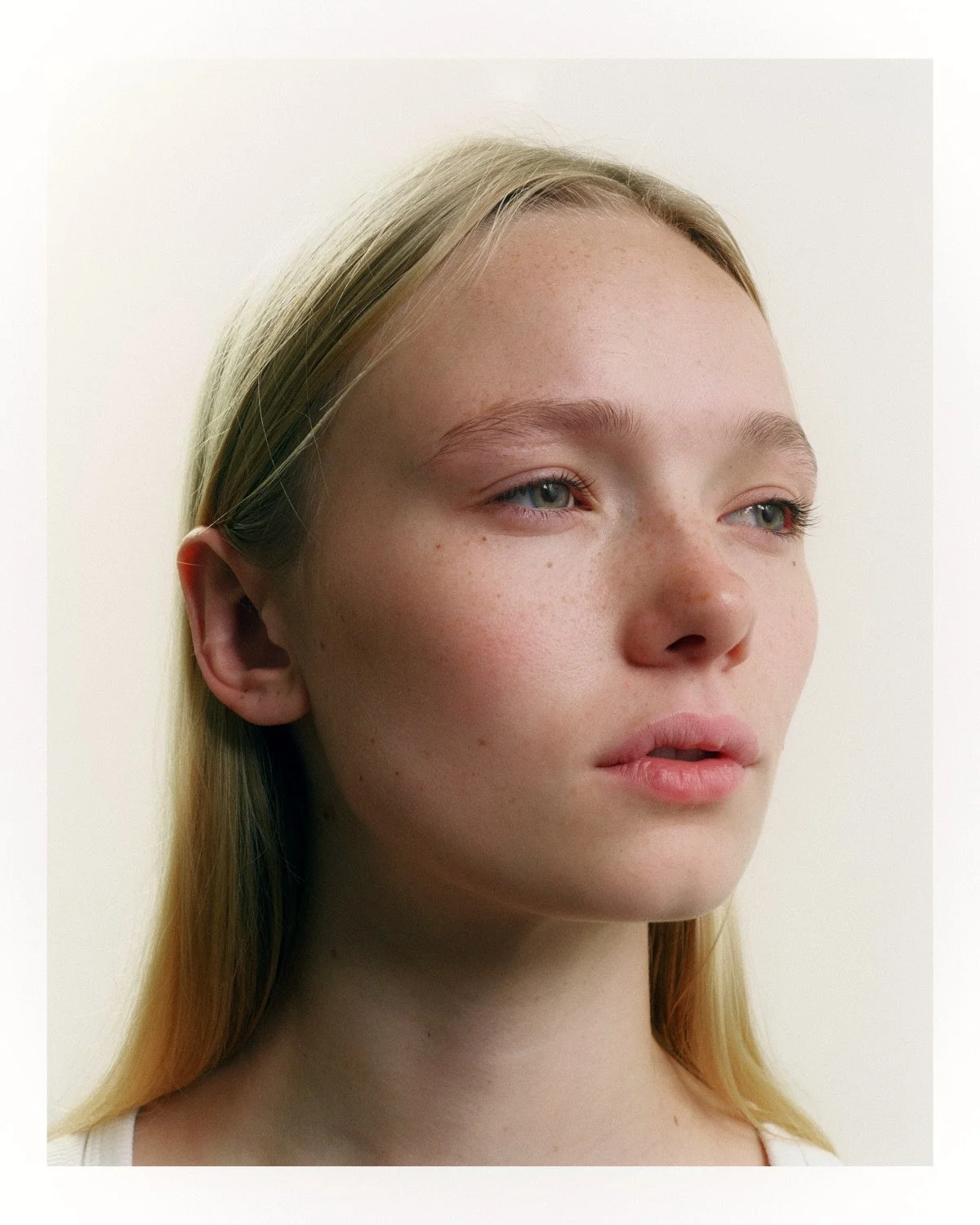 Close-up of a young woman with blonde hair and blue eyes, looking slightly to the side, against a plain light background.