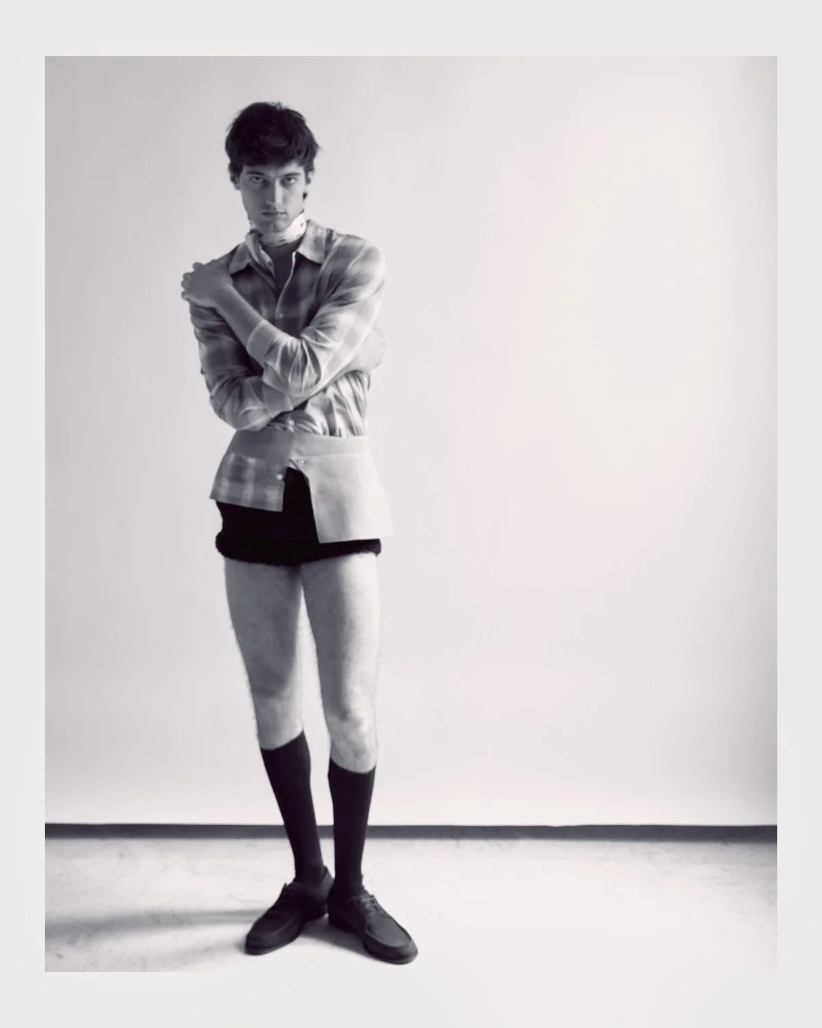 Black and white photo of a male model standing against a plain background, dressed in eclectic clothing with a layered shirt, shorts, tights, and boots, with his arms crossed and gazing at the camera.