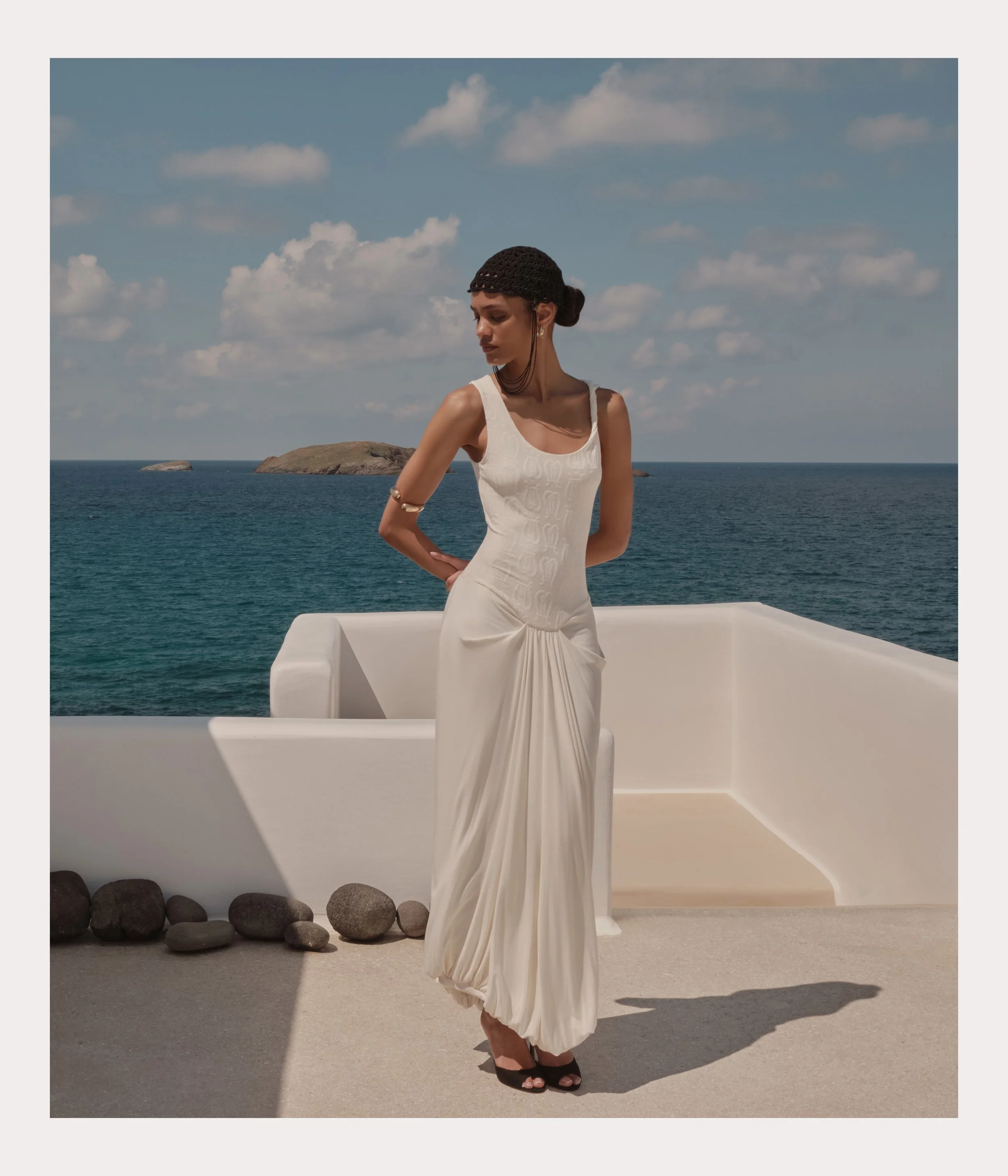 Woman in a white dress stands on a balcony overlooking the ocean, with small islands in the background and a partly cloudy sky.