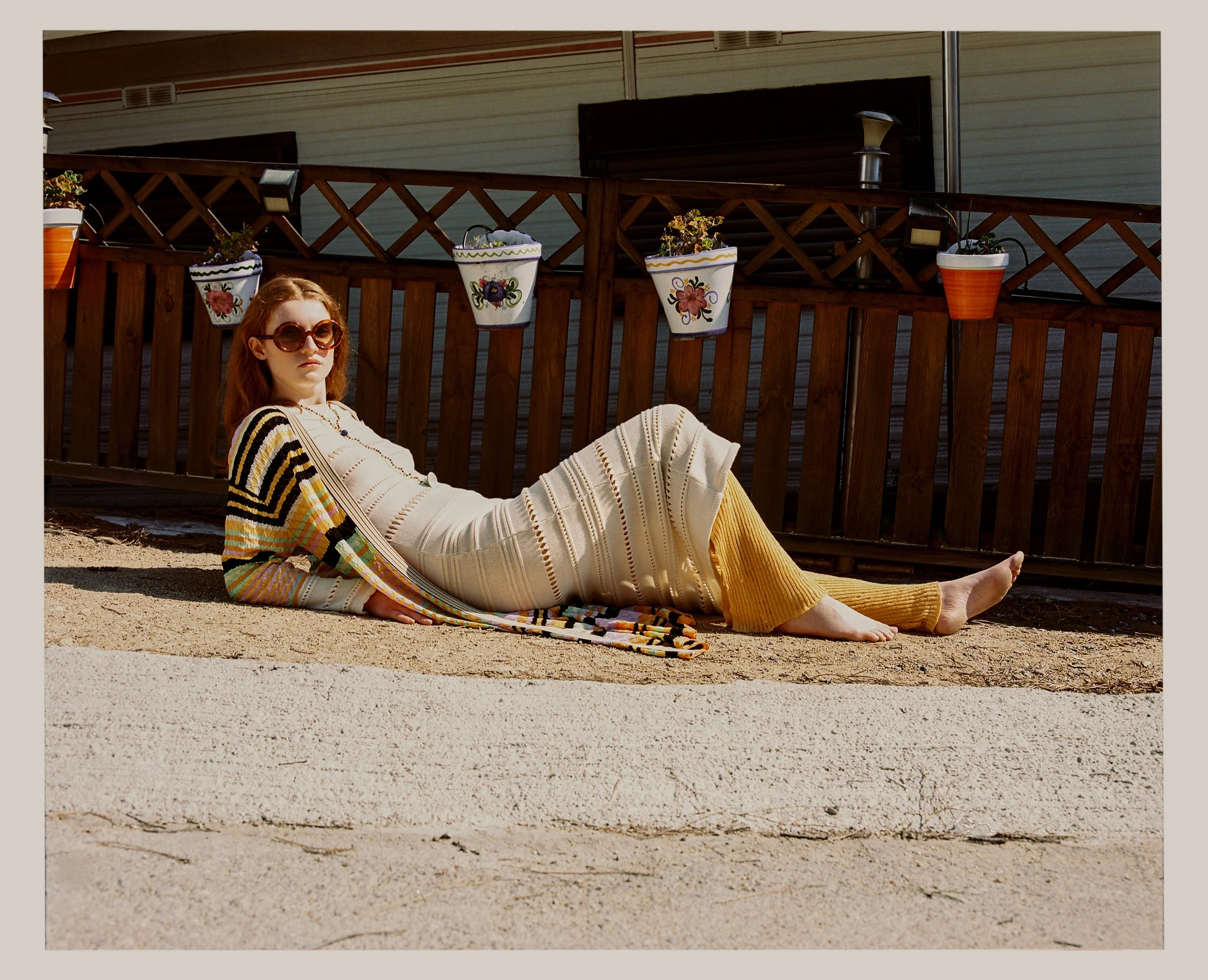 A woman with red hair wearing large sunglasses, a cream-colored knit dress, and mustard yellow pants, lying on her side on a concrete sidewalk, leaning on her elbow, in front of a wooden fence with hanging flower pots, in a sunny outdoor setting.
