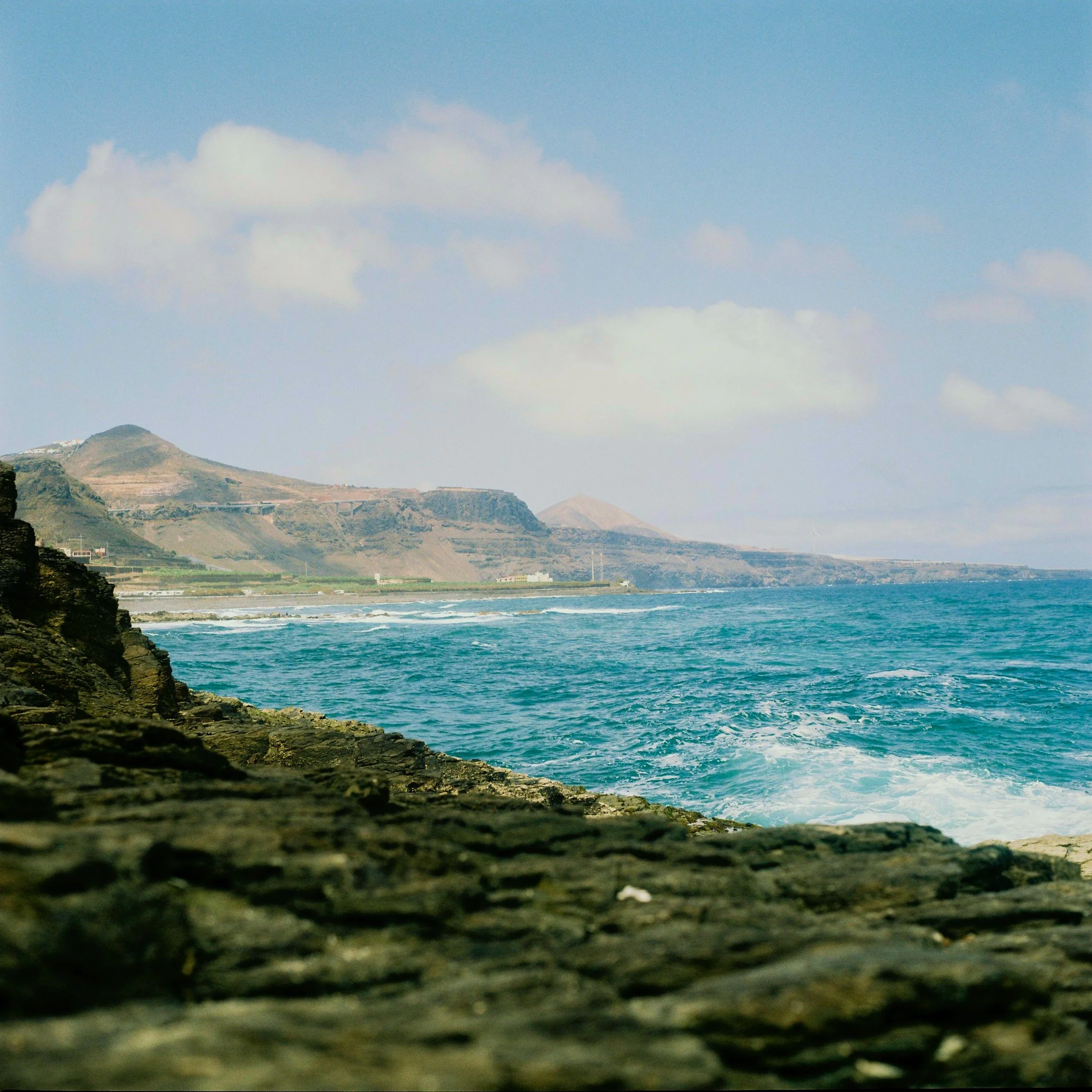 Landscape of the ocean along a rocky coastline with hills in the background and partly cloudy sky.