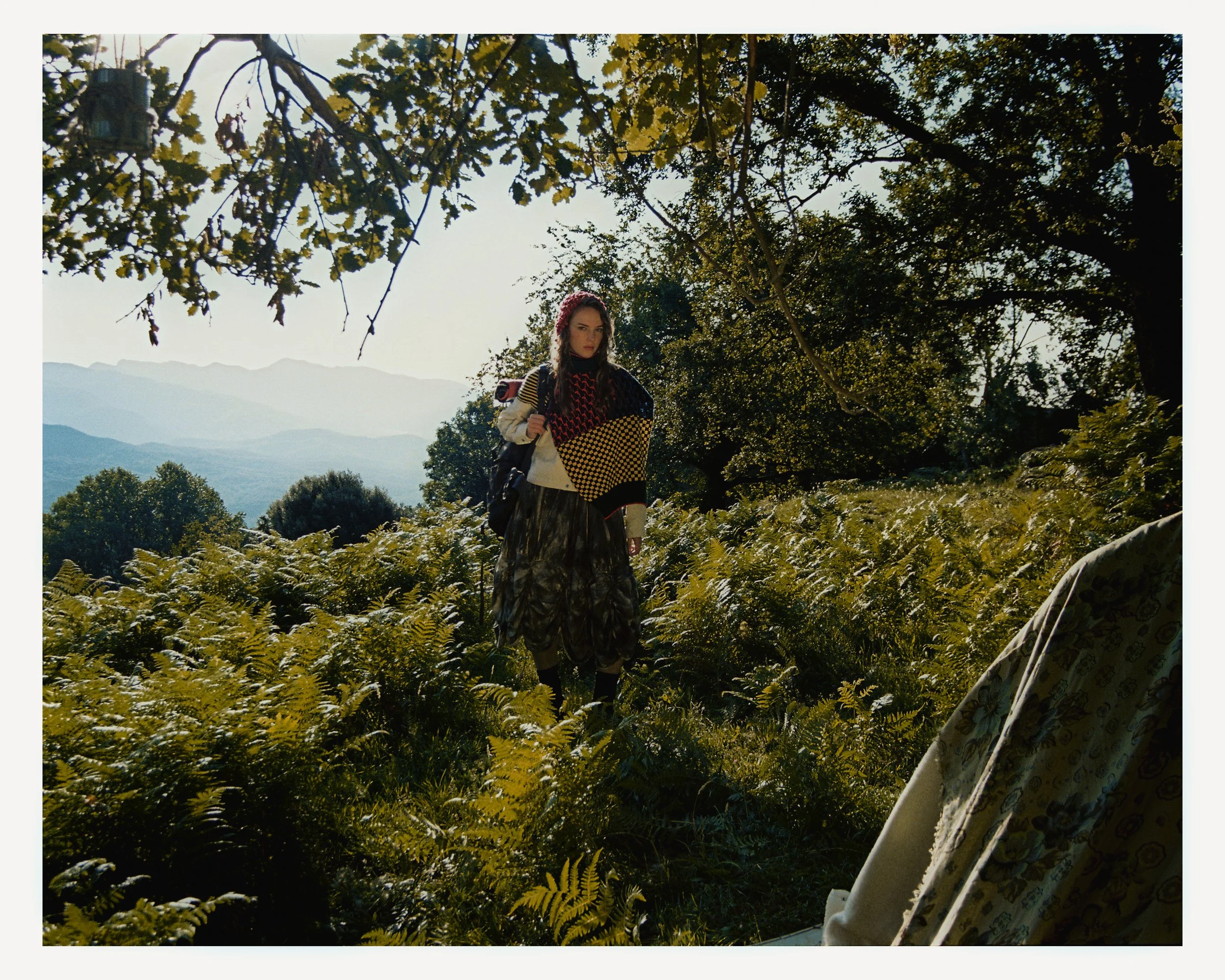 A woman stands in a lush, green forest clearing with mountains in the background. She wears a patterned poncho, long skirt, and holds a backpack, with trees and sunlit foliage around her.