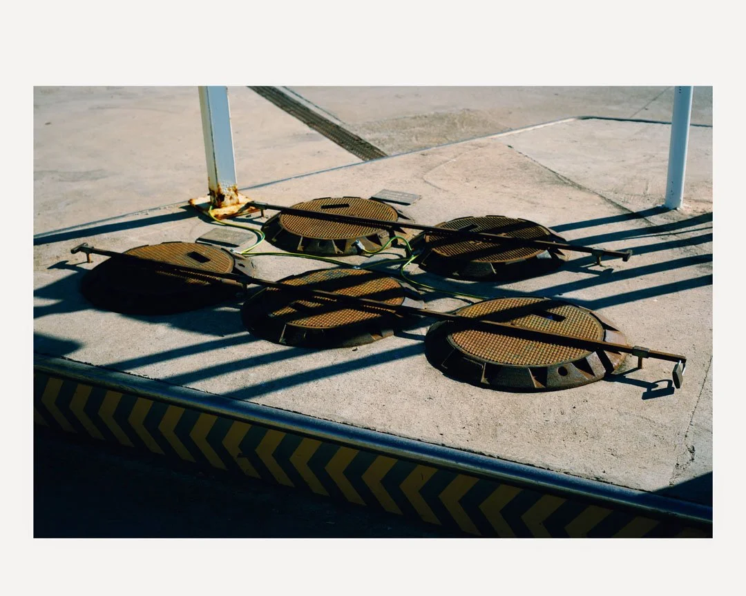 Six open manhole covers with attached tools, lying on a concrete sidewalk next to a caution barrier at a construction site.