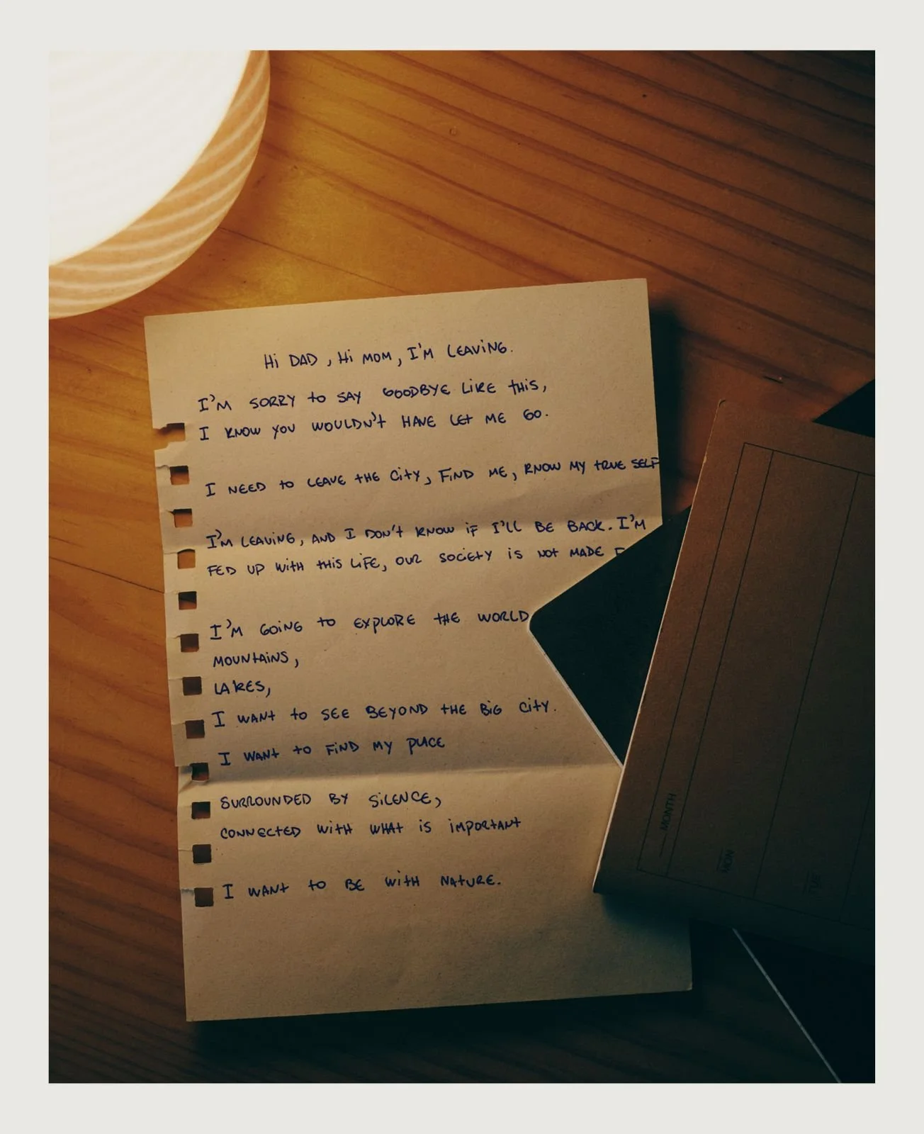 A handwritten note on torn notebook paper with a message about leaving home, placed on a wooden surface near a dark notebook and a lamp.