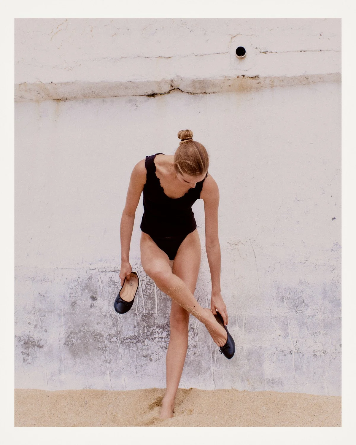 A woman in a black swimsuit, holding a pair of black shoes, stands outdoors on sand against a weathered white wall, lifting her leg and looking down.