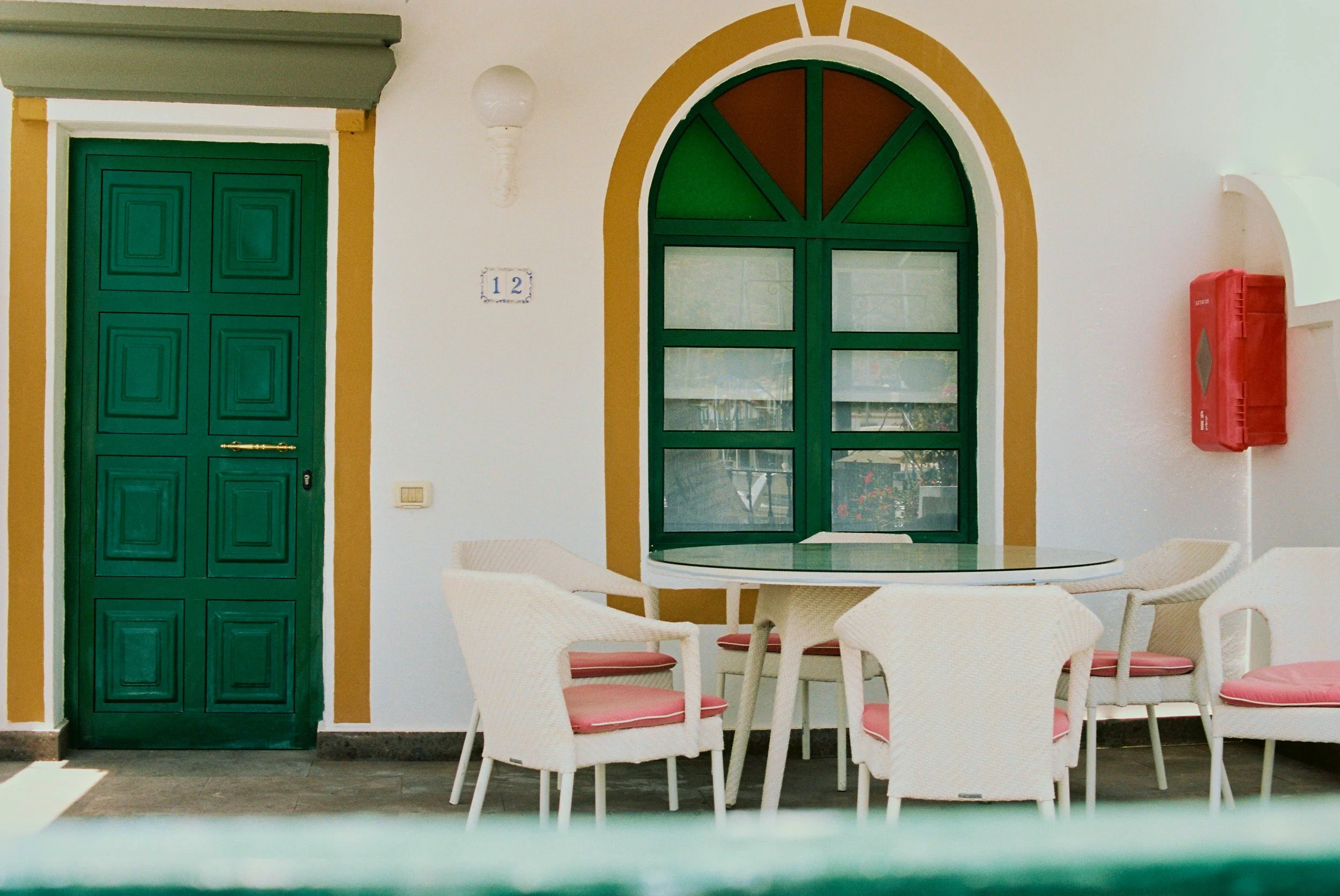 A porch area with a green wooden door, a colorful arched window, a round table, and four white chairs with pink cushions. There is a white wall, a wall-mounted light, a house number 12, and a red fire extinguisher box.
