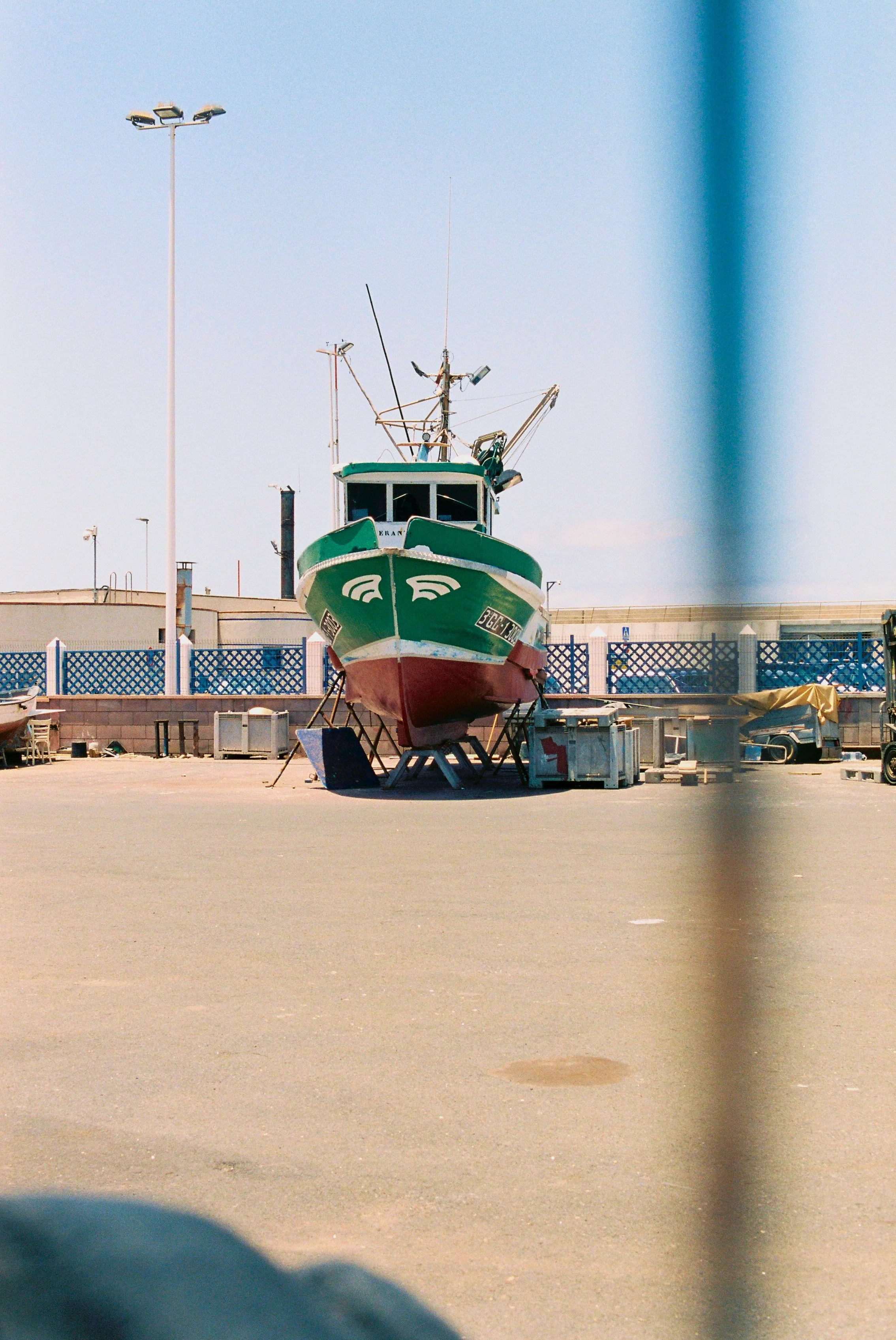 A green and red fishing boat on boat stands in a boatyard, with equipment and a fence in the background.