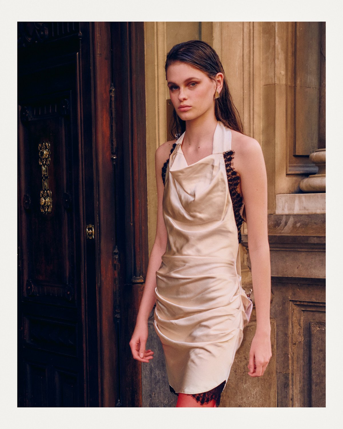 A young woman with long, dark hair wearing a sleeveless, satin beige dress with black lace side panels, standing in front of a wooden door and stone wall.