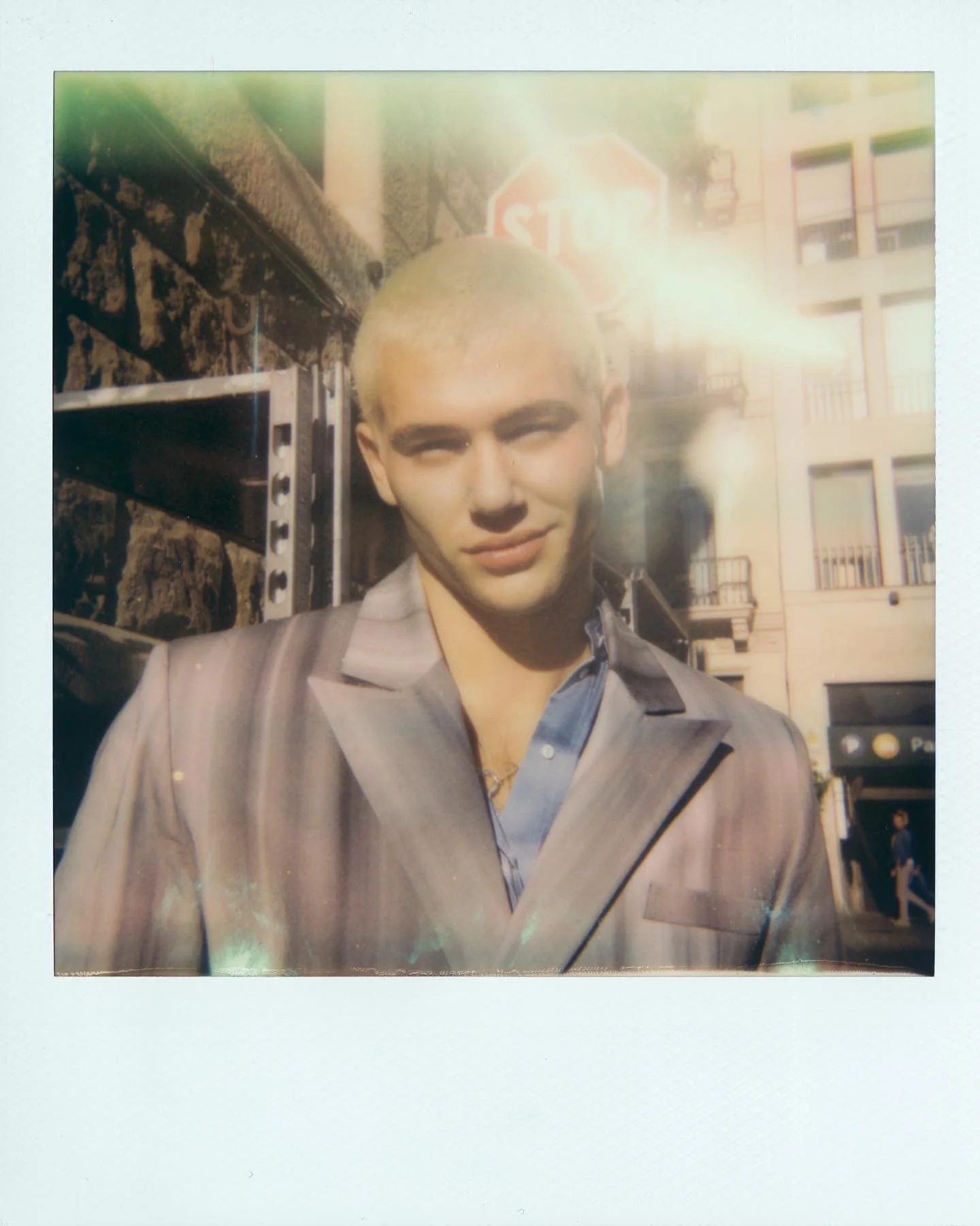 A young man with short blonde hair is standing outdoors in front of a building and a stop sign, with sunlight shining brightly behind him.