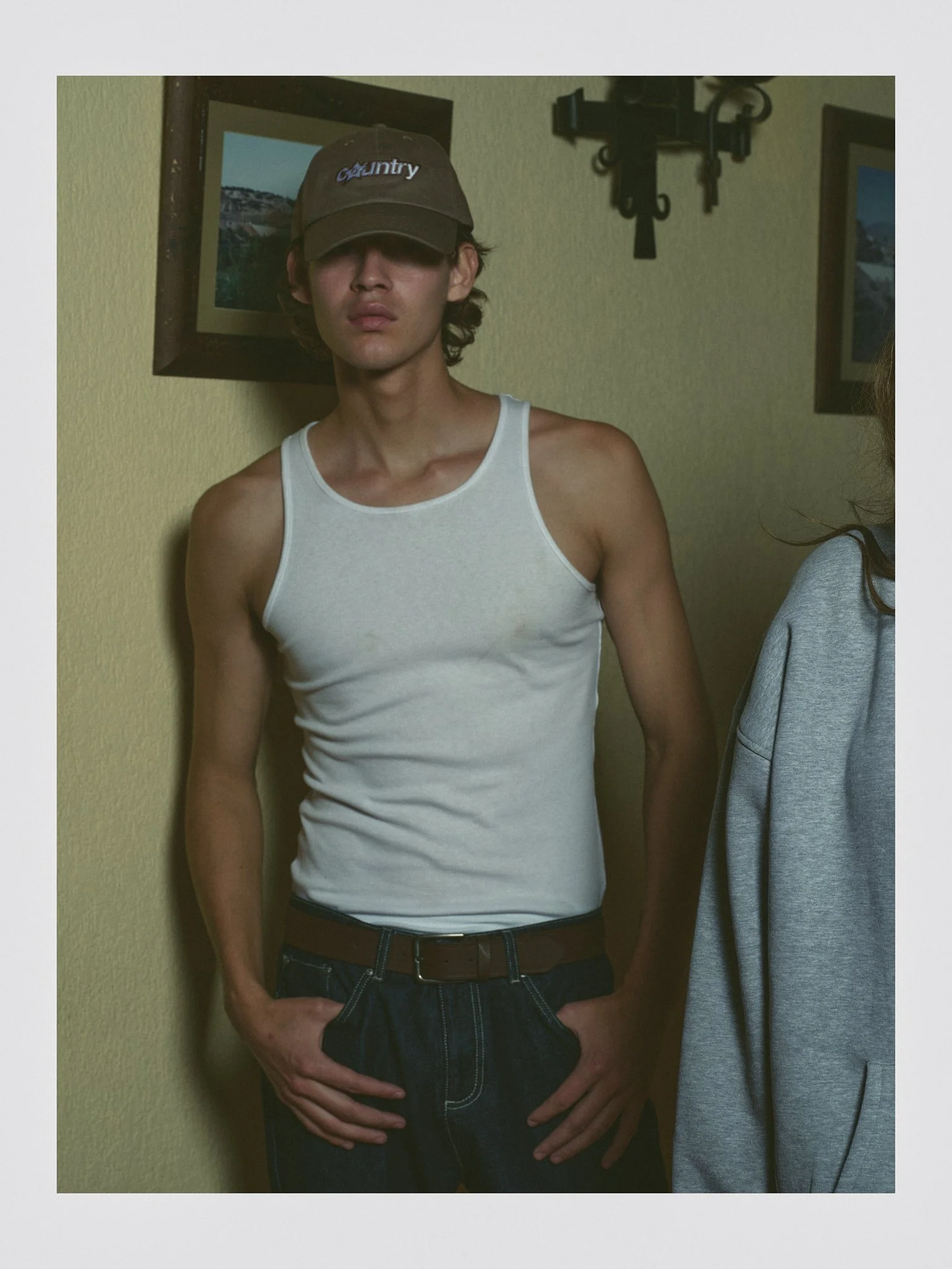 Young man with long hair wearing a dark cap, white tank top, unbuttoned jeans, and a brown belt, standing against a yellow wall with framed pictures and a coat hanging.