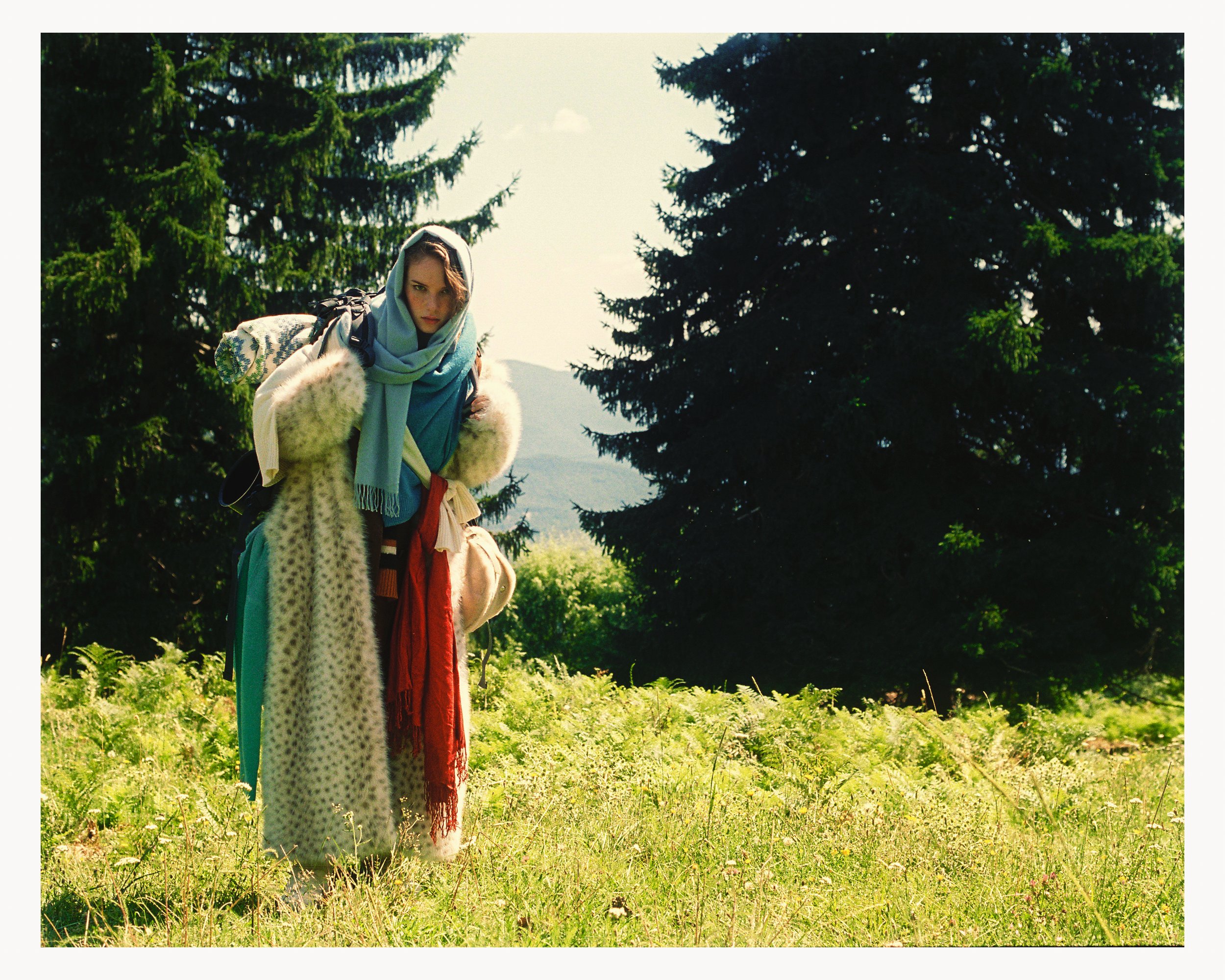 A woman dressed warmly with a scarf over her head and a long coat, standing in a grassy field with pine trees and mountains in the background.