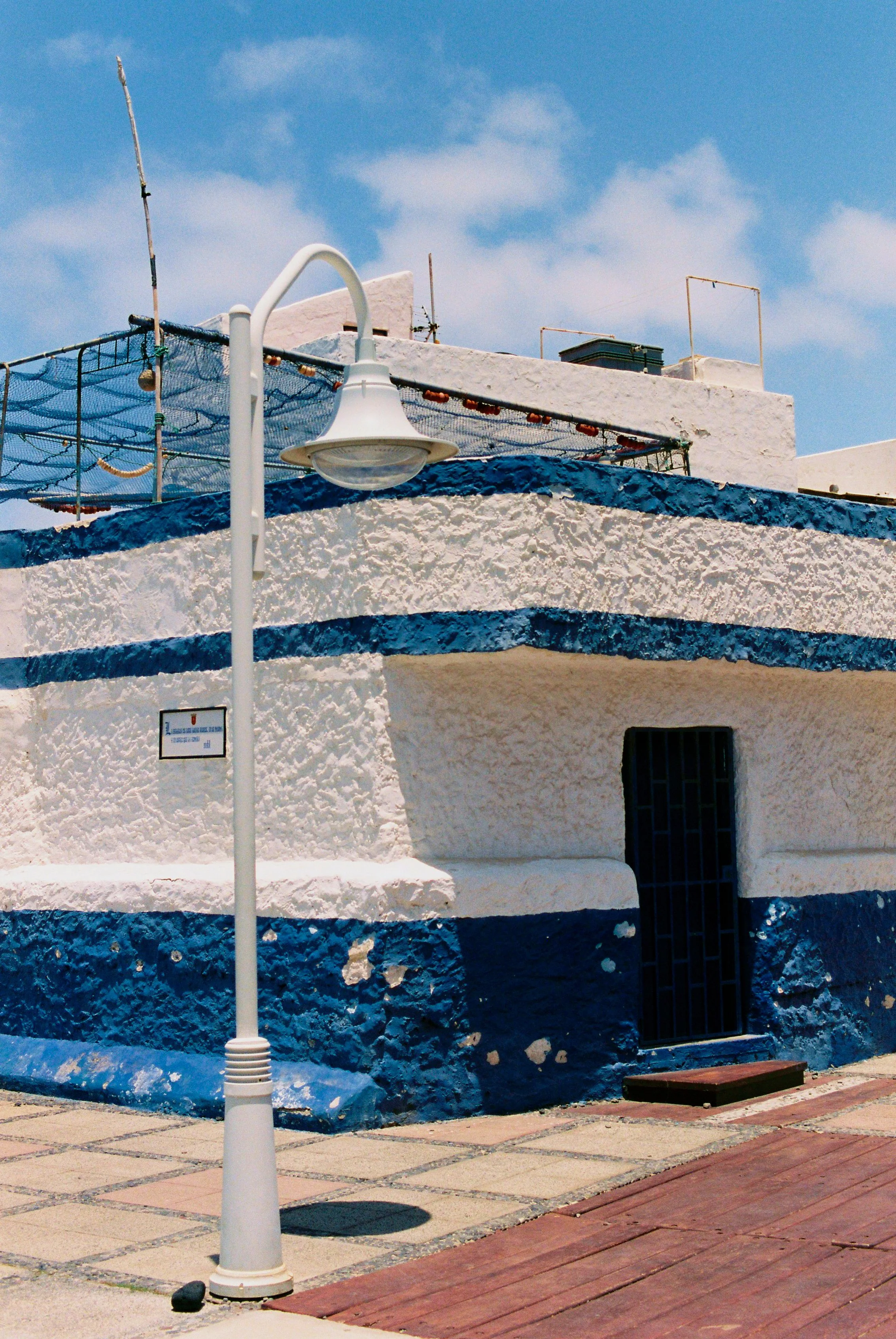 A white and blue building with a textured exterior, a black door, and a street lamp in the foreground. The building has a rooftop area with some antennas and a clear blue sky overhead.