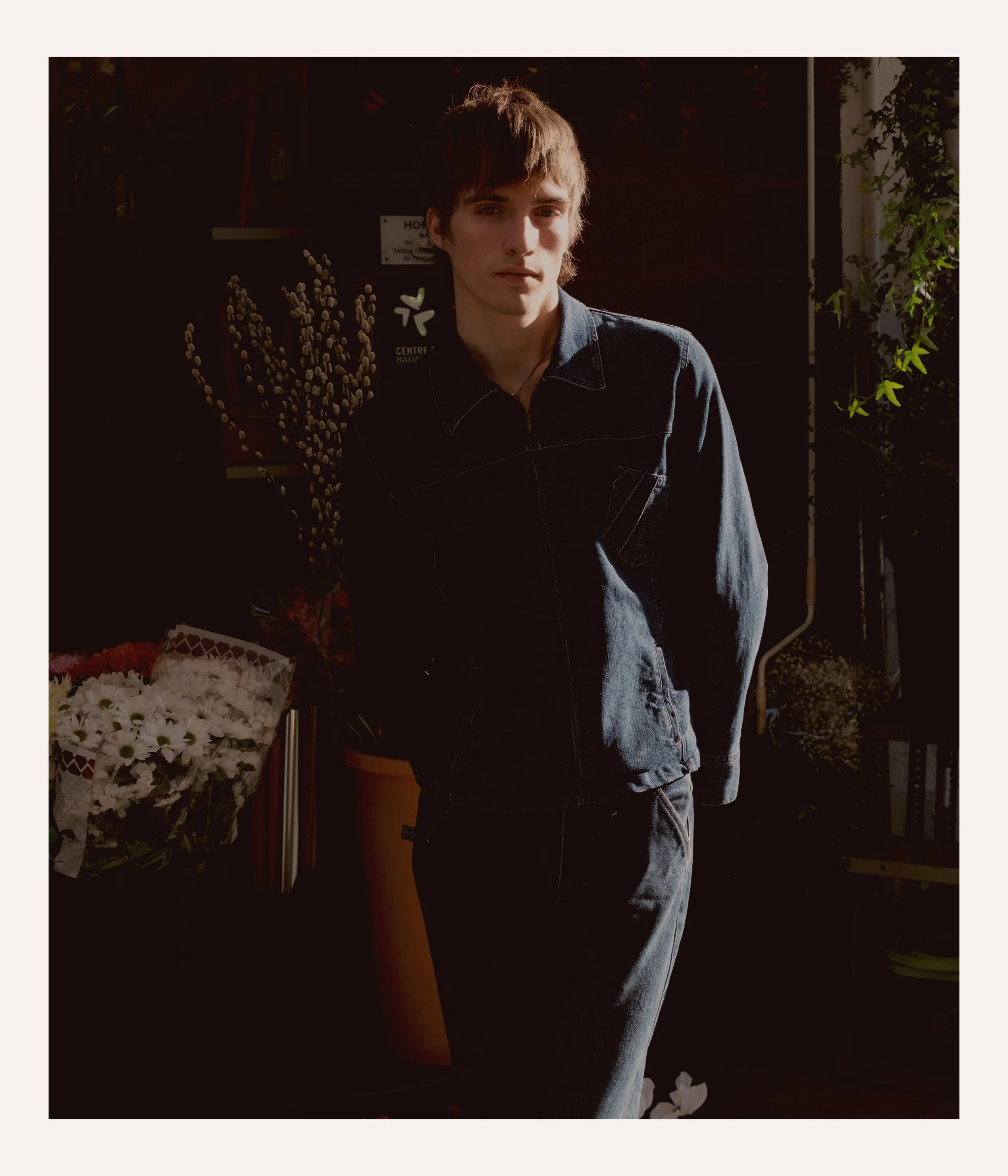 Young man in dark denim jacket and pants standing in a dimly lit room with plants and flowers around him.