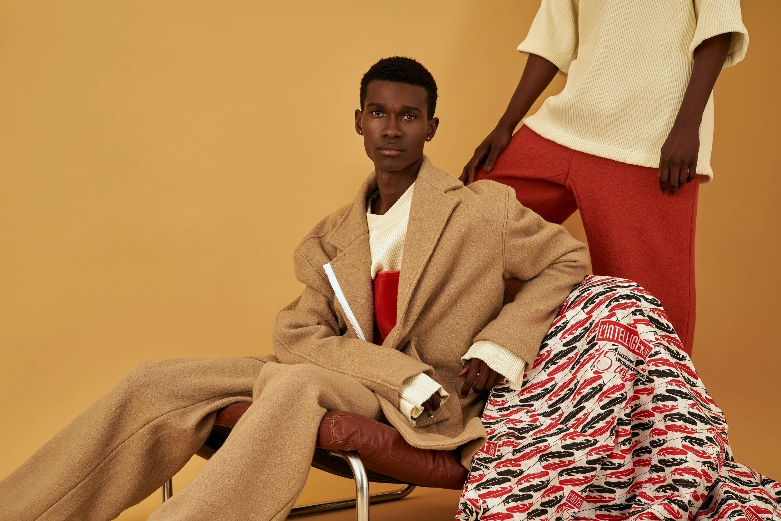 A young person with dark skin and short curly hair, dressed in a beige coat over a cream sweater with red details, sitting on a chair against a solid yellow background. Another person, standing to their side, wears a cream sweater and red pants. Ther