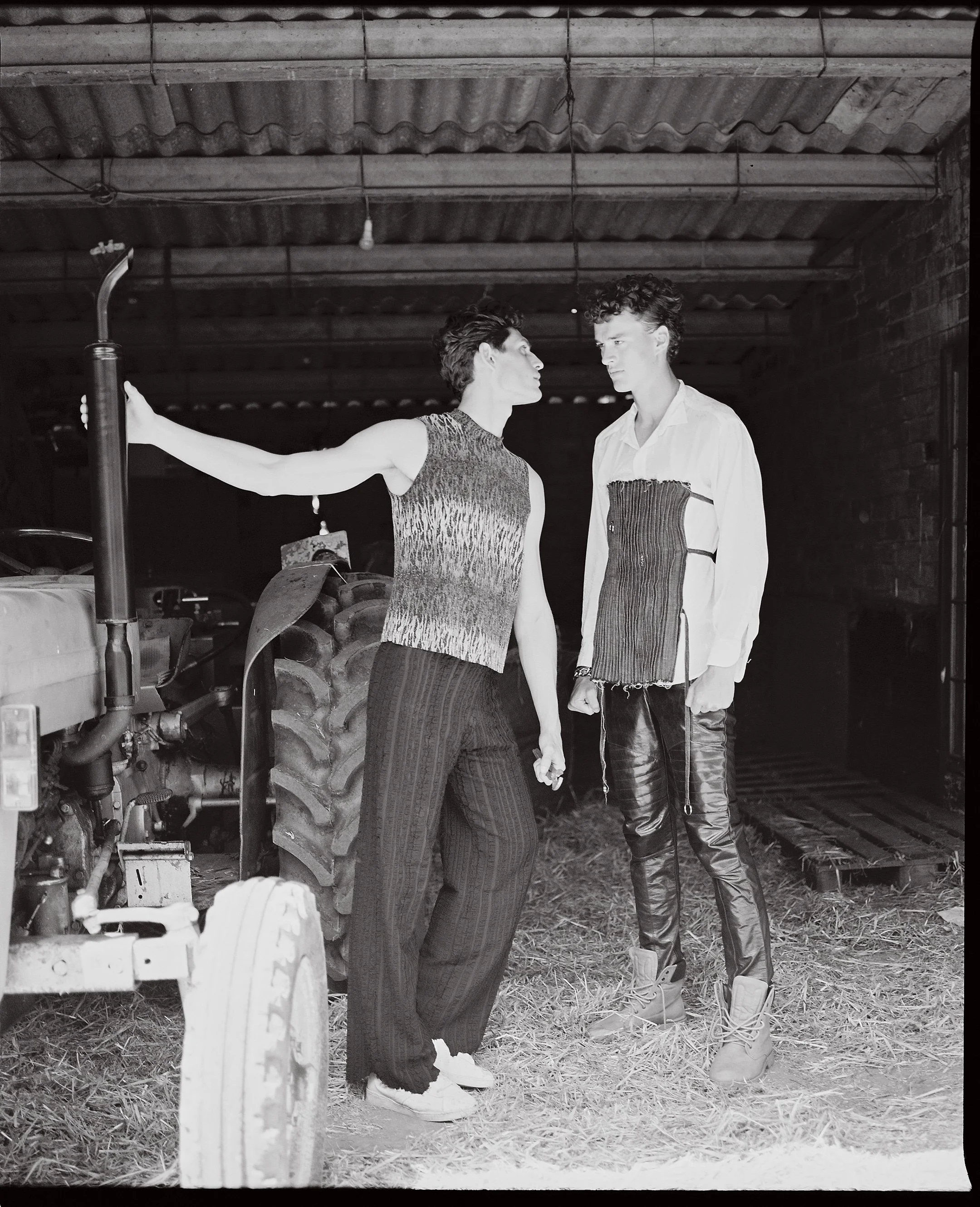 Two young people stand close in a barn-like setting, with one reaching out and leaning toward the other. Both wear distinctive fashion-forward clothing, and a large tractor is visible on the left side.