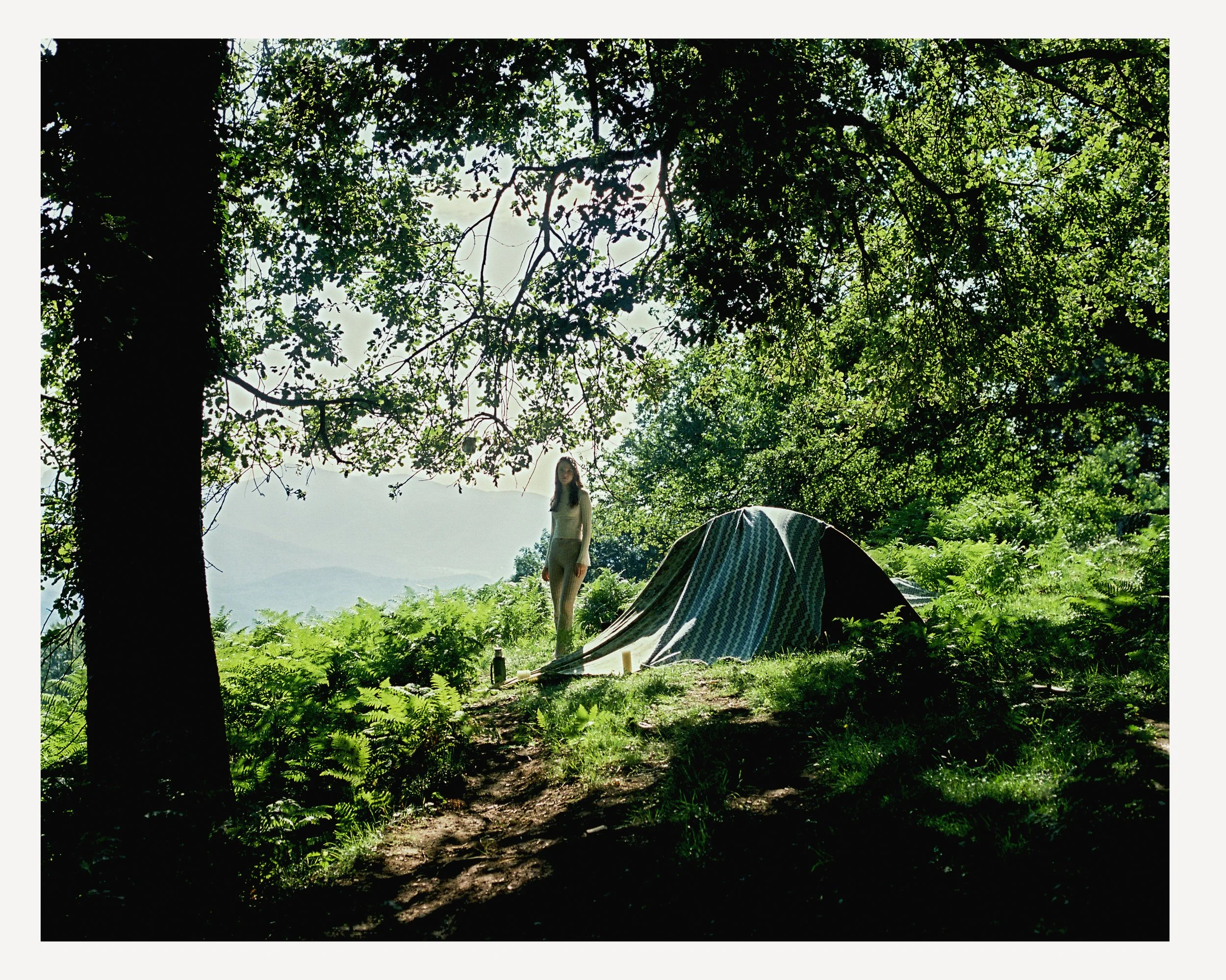 A person standing near a tent in a lush, green forest with large trees and ferns, with mountains visible in the background under a partly cloudy sky.