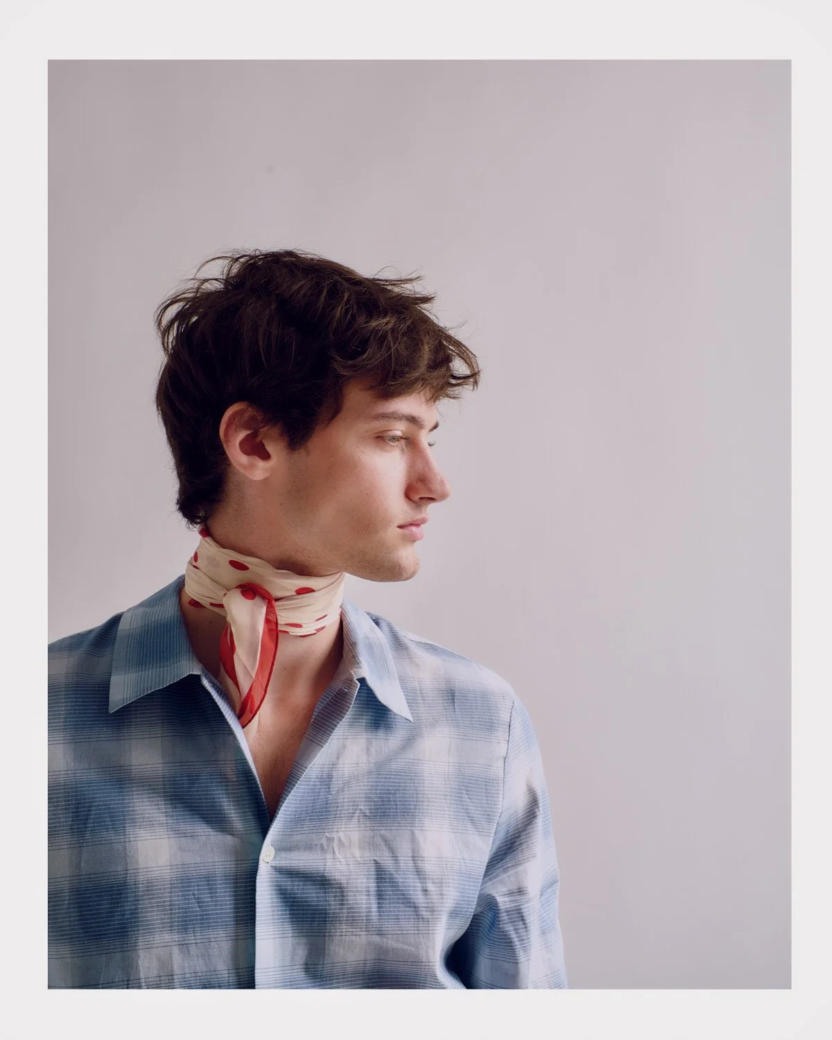 A young man with brown hair wearing a blue and white plaid shirt and a white scarf with red polka dots around his neck, looking to the right.