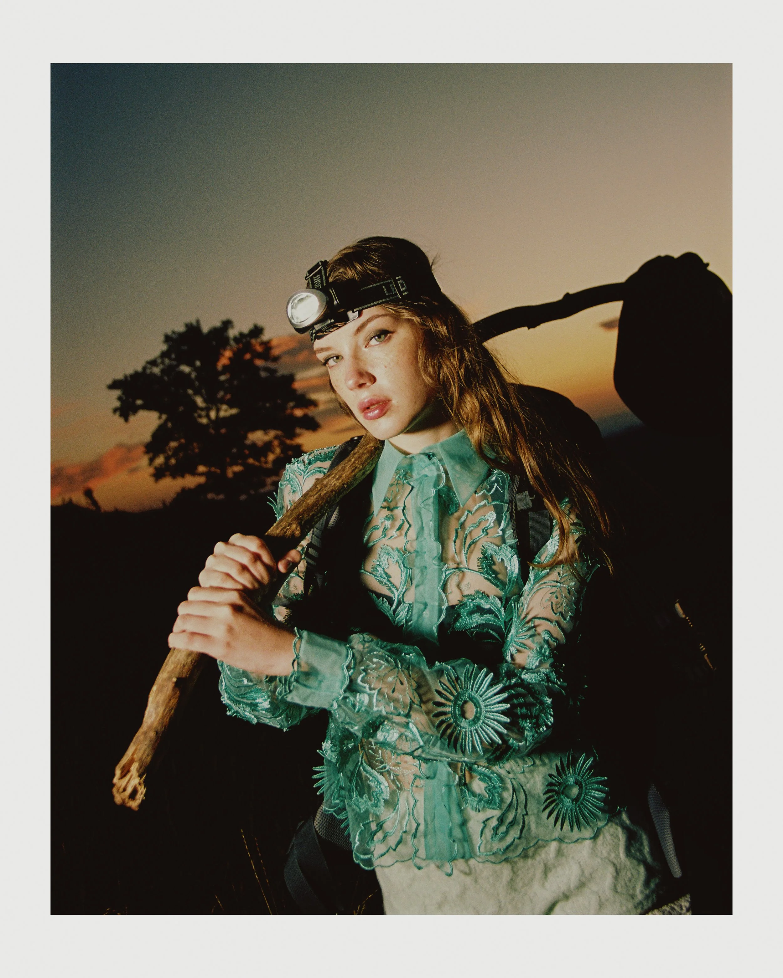 Young woman with long hair wearing a headlamp and a sheer embroidered blouse, holding a walking stick, at sunset in a natural outdoor setting.