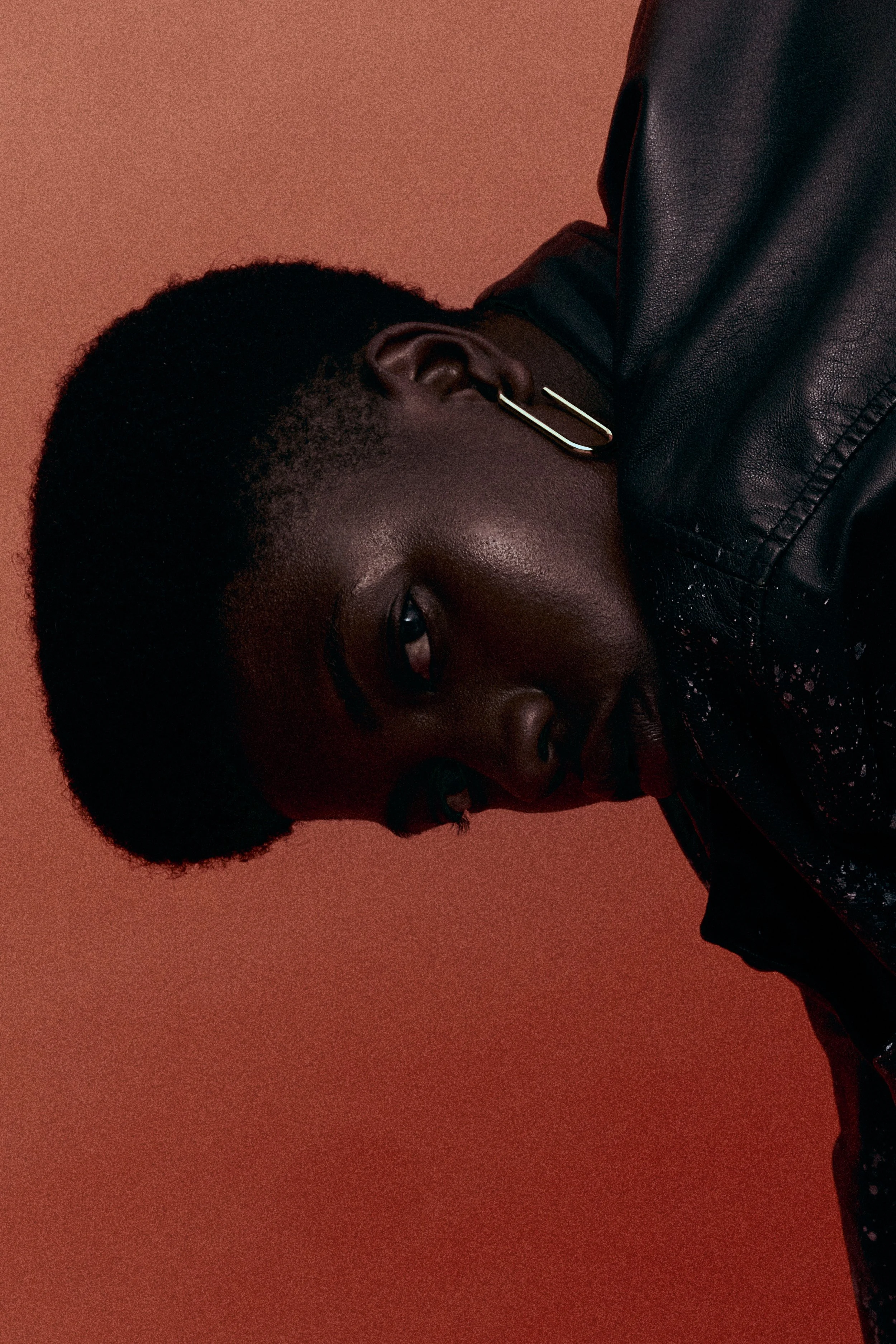 Portrait of a person with dark skin and a short, cropped hairstyle, wearing a black leather jacket and a gold earring, posed against a brownish-orange background.