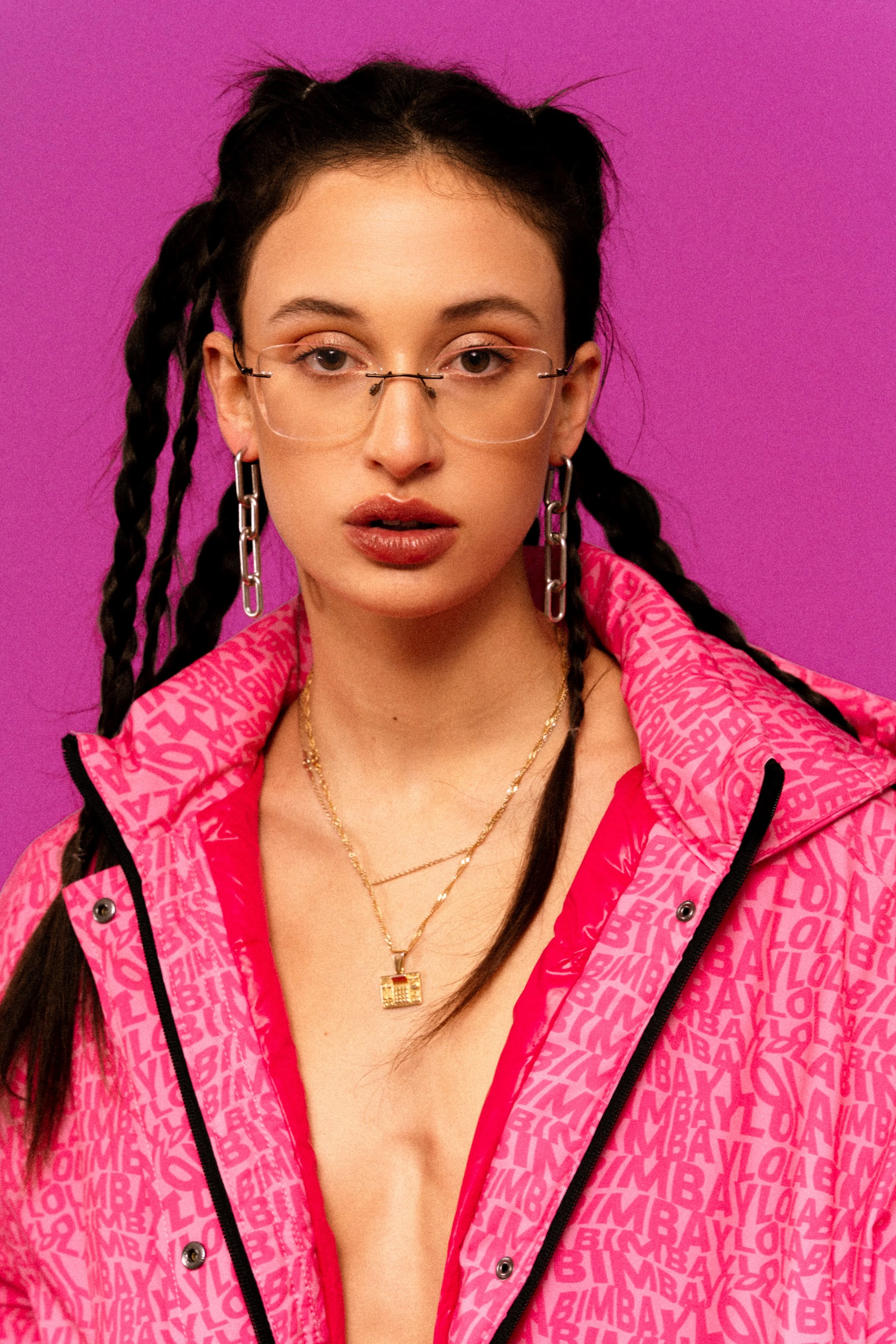 Portrait of a young woman with dark braided hair, wearing glasses, large chain earrings, layered gold necklaces, and a pink jacket with text pattern, standing against a purple background.