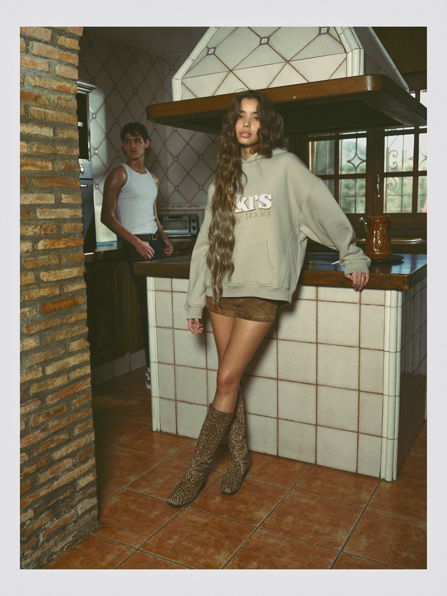 Young woman with long wavy hair wearing a beige hoodie with logo, brown shorts, and leopard print boots standing in a rustic kitchen, with a young man in a white tank top standing in the background near a counter.