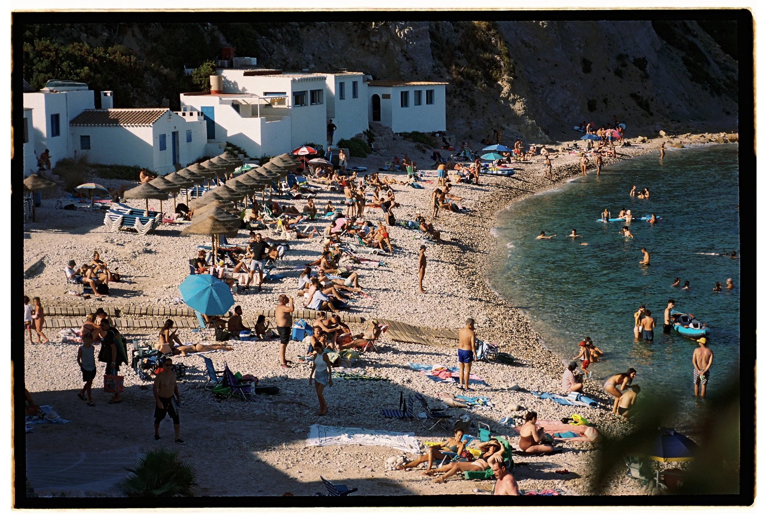 People relaxing on a crowded beach with umbrellas and lounge chairs, some swimming and wading in the water, white buildings on a rocky cliff in the background.