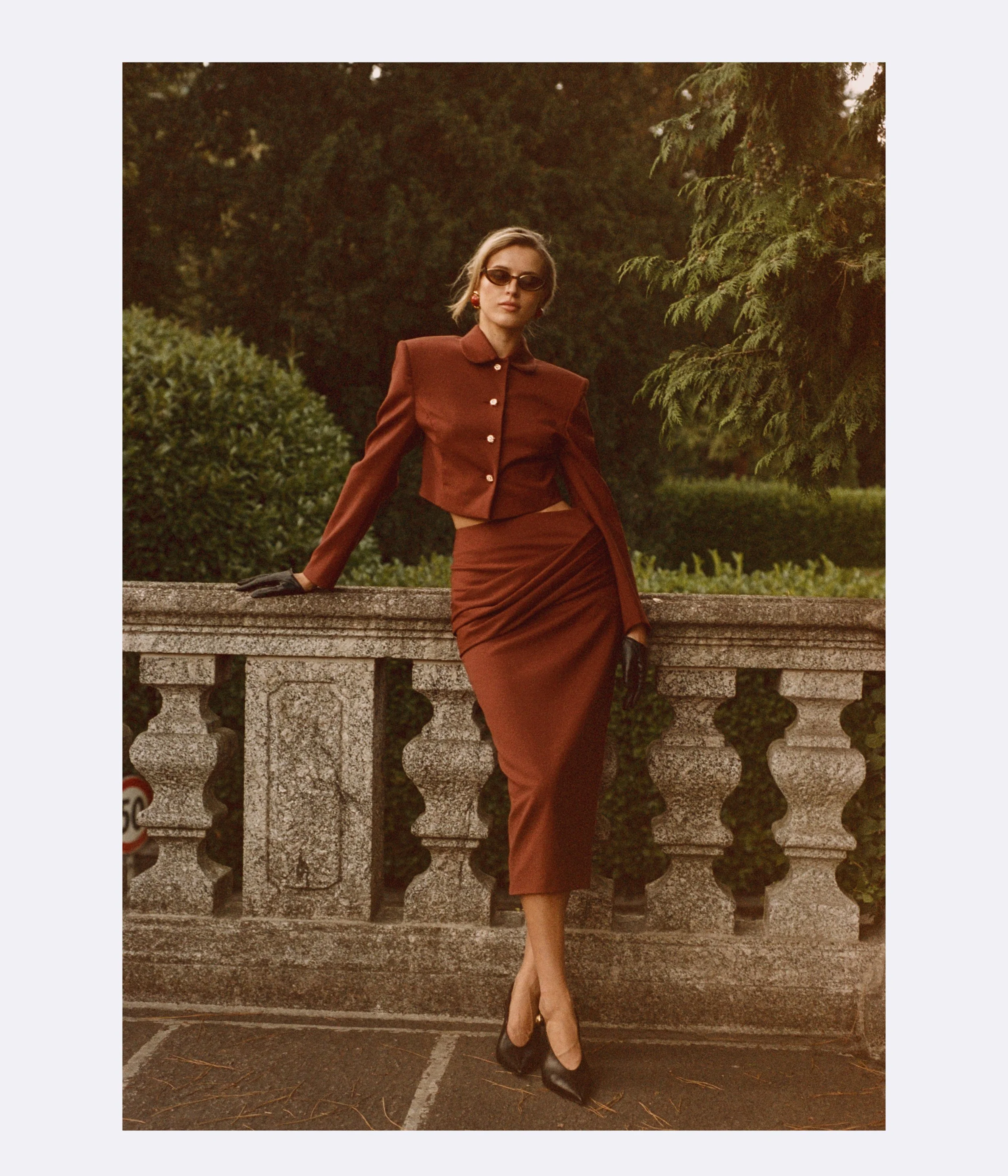A woman in a vintage brown outfit with a matching jacket and skirt, wearing black gloves, high heels, and sunglasses, sitting on a stone railing in an outdoor park or garden setting during sunset.