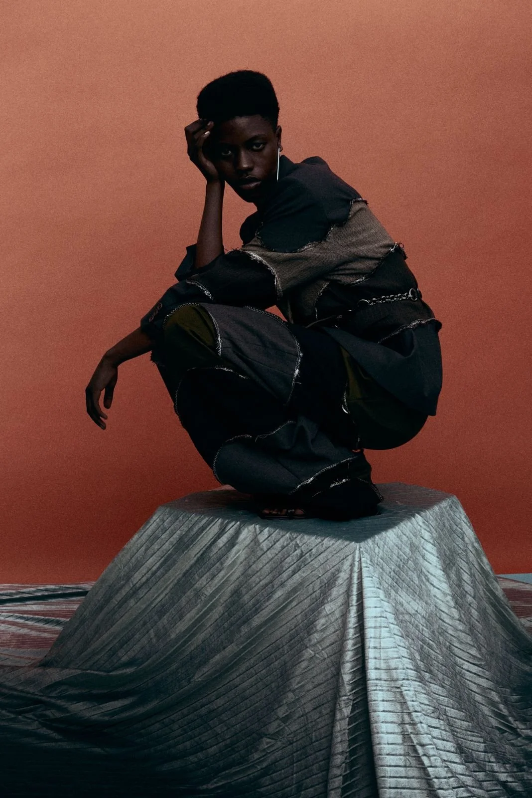 A person crouching on a metallic platform against a brown background, wearing a black and metallic patchwork outfit and an earring.