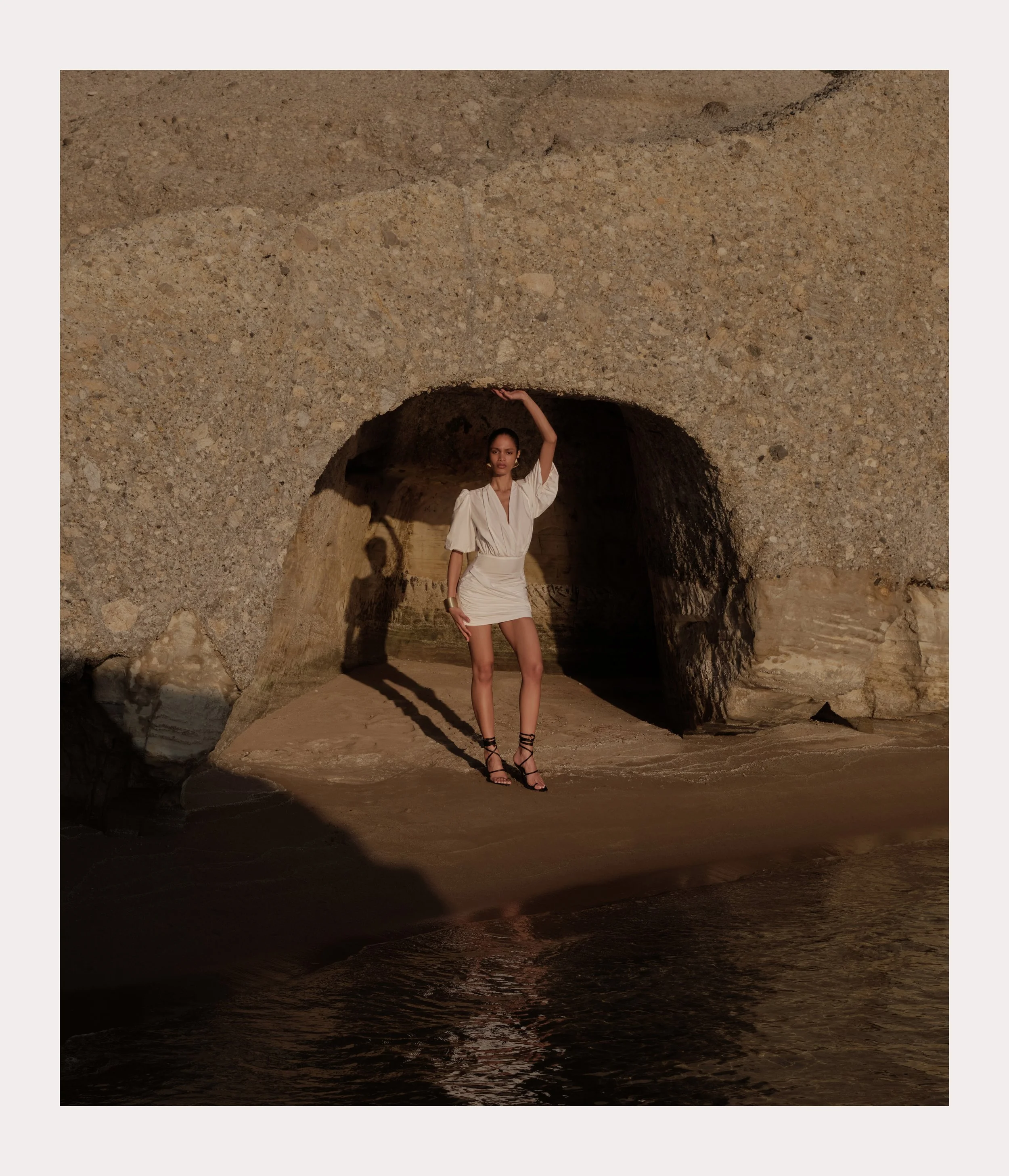 A woman in a white dress and black strappy heels standing on a sandy beach inside a cave, with part of her shadow cast on the rock wall behind her.