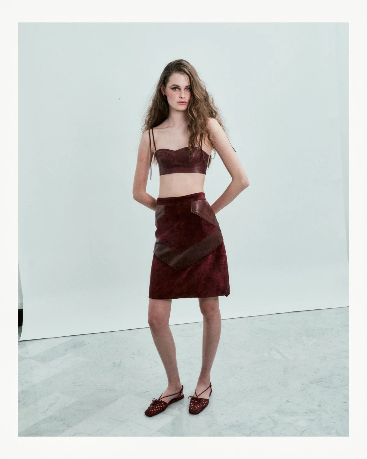A woman with long wavy hair wearing a burgundy crop top and matching skirt, standing against a plain white backdrop.