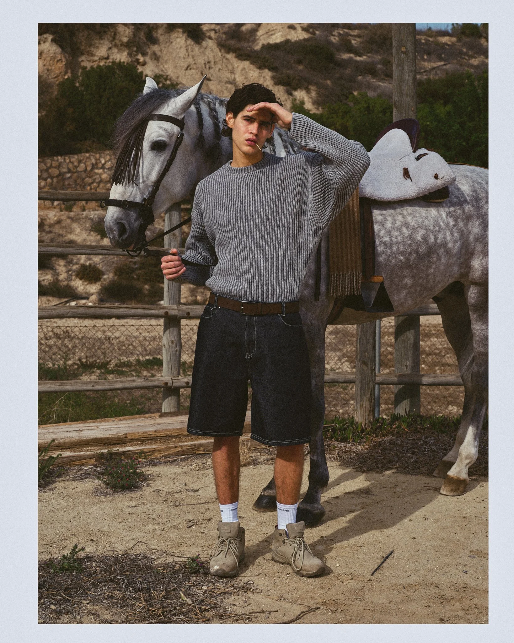 A young man stands outdoors holding a lead rope, with a gray horse behind him, in front of a wooden fence and hillside. The man wears a gray striped sweater, black shorts, white socks, and tan shoes, and shades his eyes from the sun with his hand.