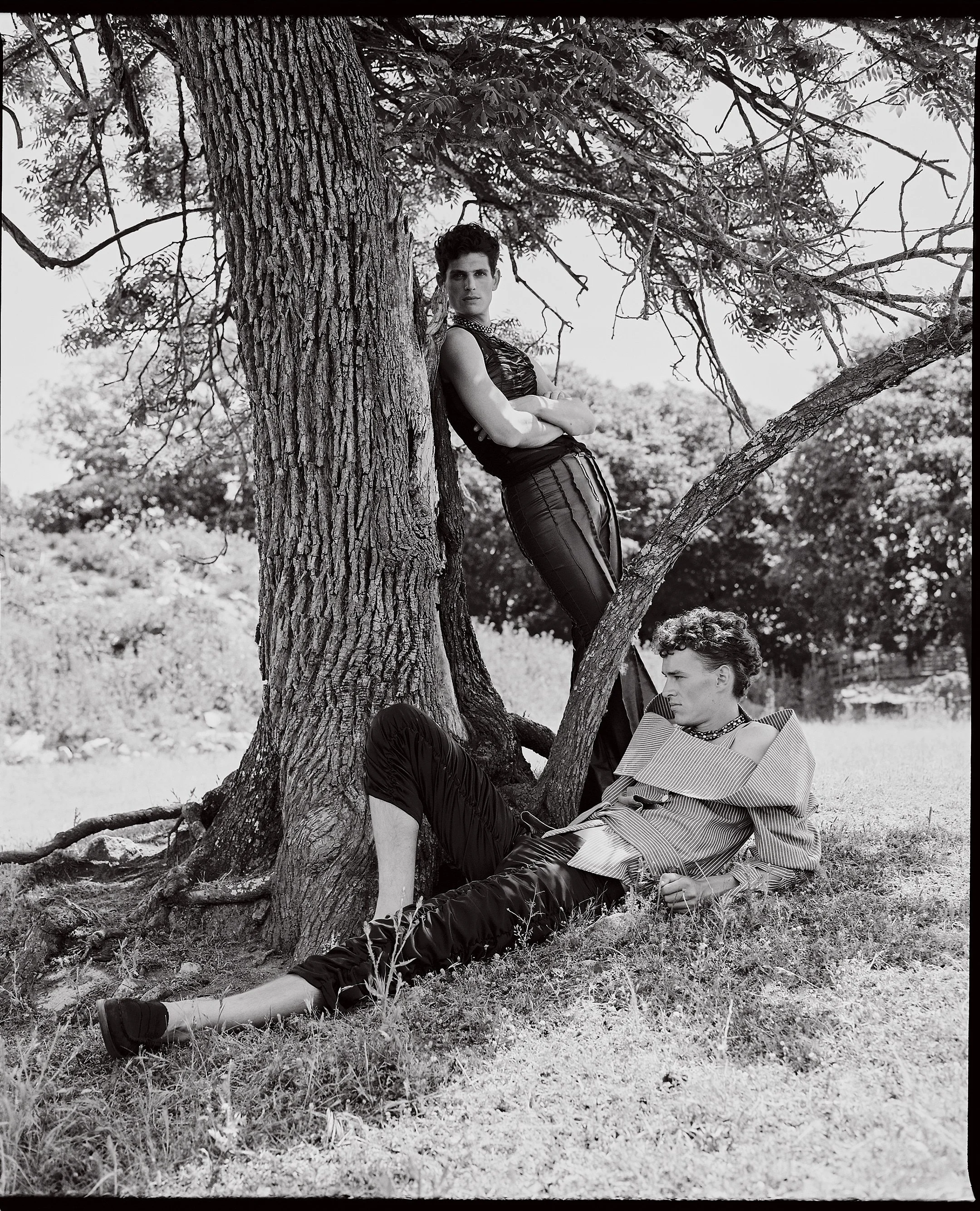 Two androgynous models posing outdoors under a large tree. One model is leaning against the trunk with arms crossed, while the other is reclining on the ground with one leg bent. Both are dressed in fashion-forward clothing.