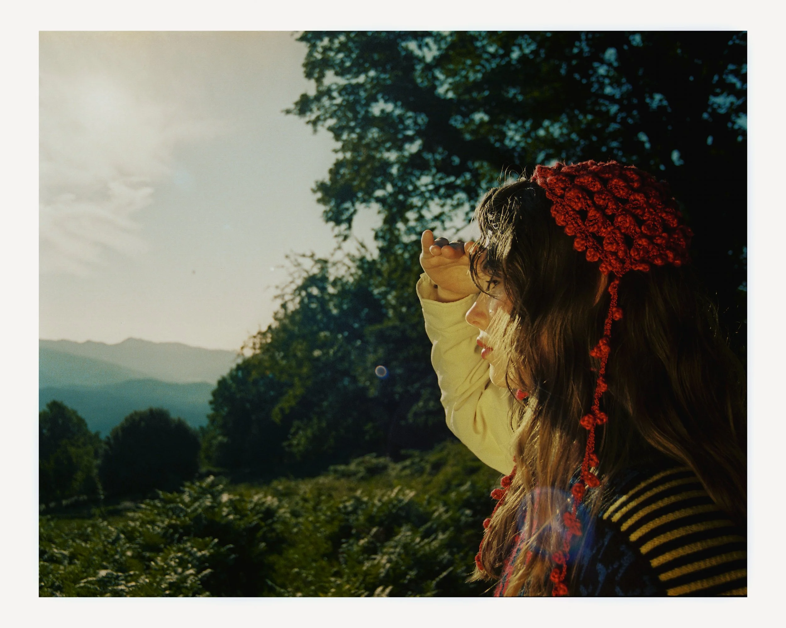 A woman wears a red hat with pom-poms, standing outdoors in a natural landscape with trees and mountains, gazing into the distance as sunlight shines from the left.