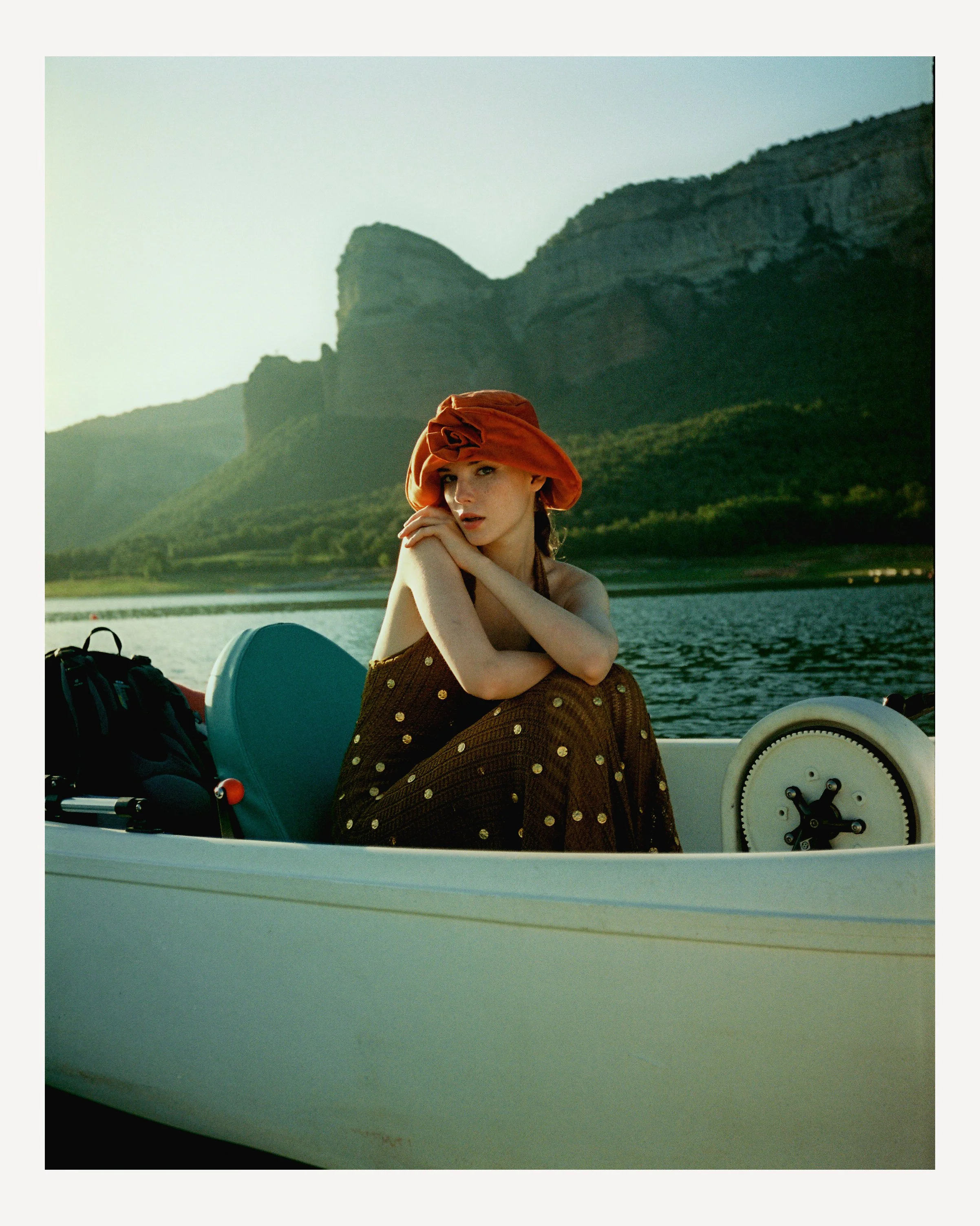 A woman wearing a brown dress with yellow polka dots and a red hat sits in a small boat on a lake with mountains in the background.