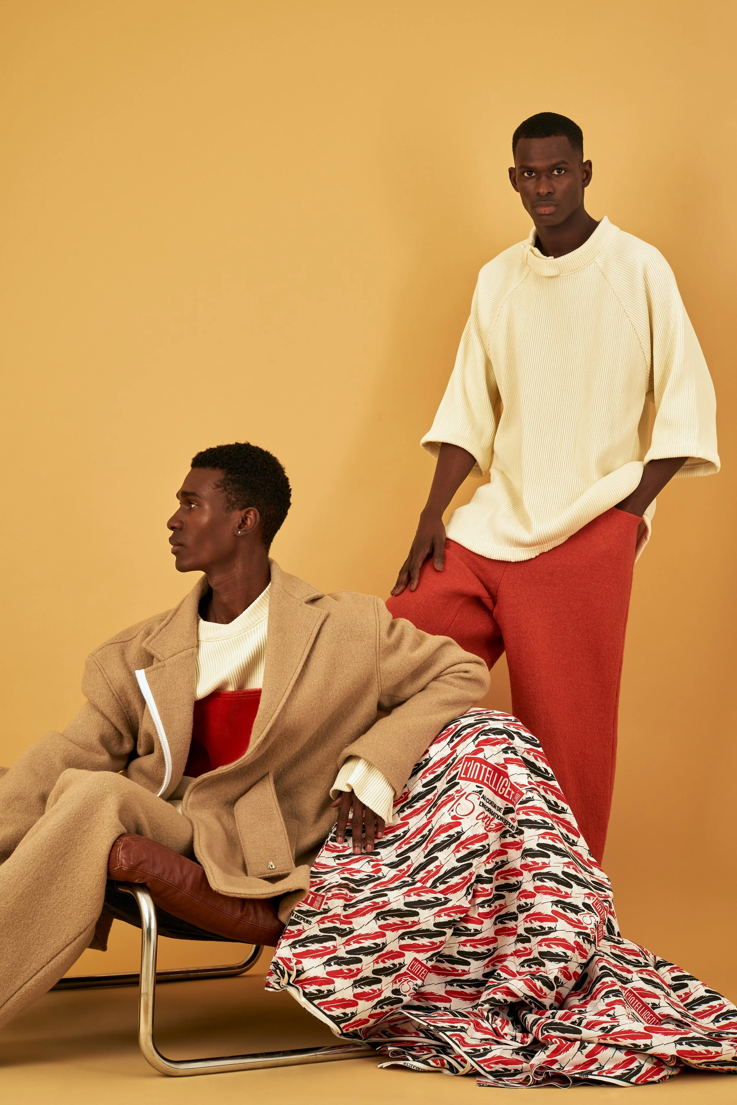 Two male models with dark skin in fashion-forward outfits posed against a yellow background. One is sitting on a chair wearing a tan coat, cream sweater, and red top. The other is standing, wearing a cream oversized sweater and red pants. A patterned