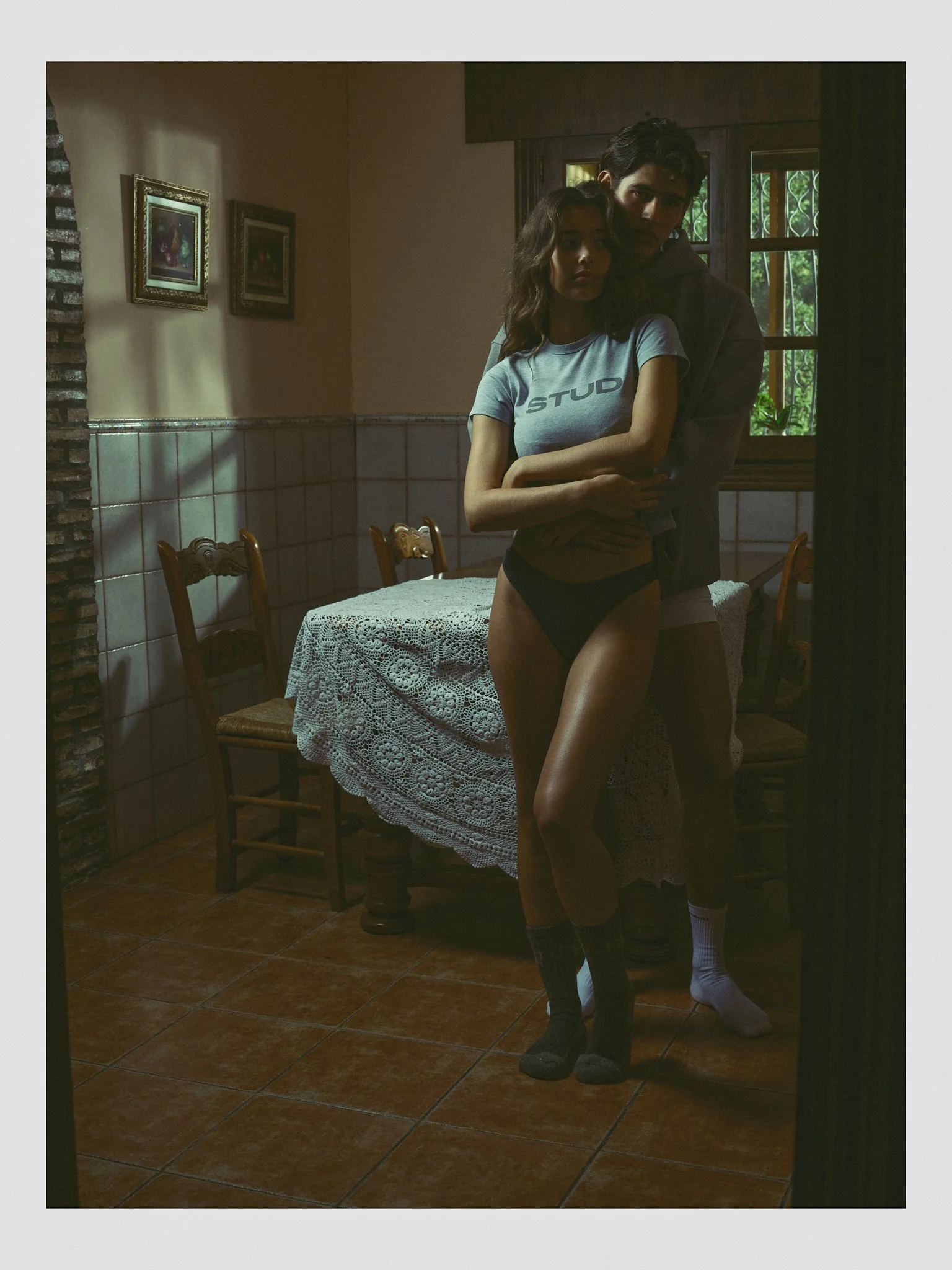 A young woman in a gray t-shirt with the word 'STUD' on it and black underwear stands with her arms crossed in front of a young man in a dark jacket and shorts. They are in a dimly lit room with a tiled wall, wooden chairs, a table with a lace tablec