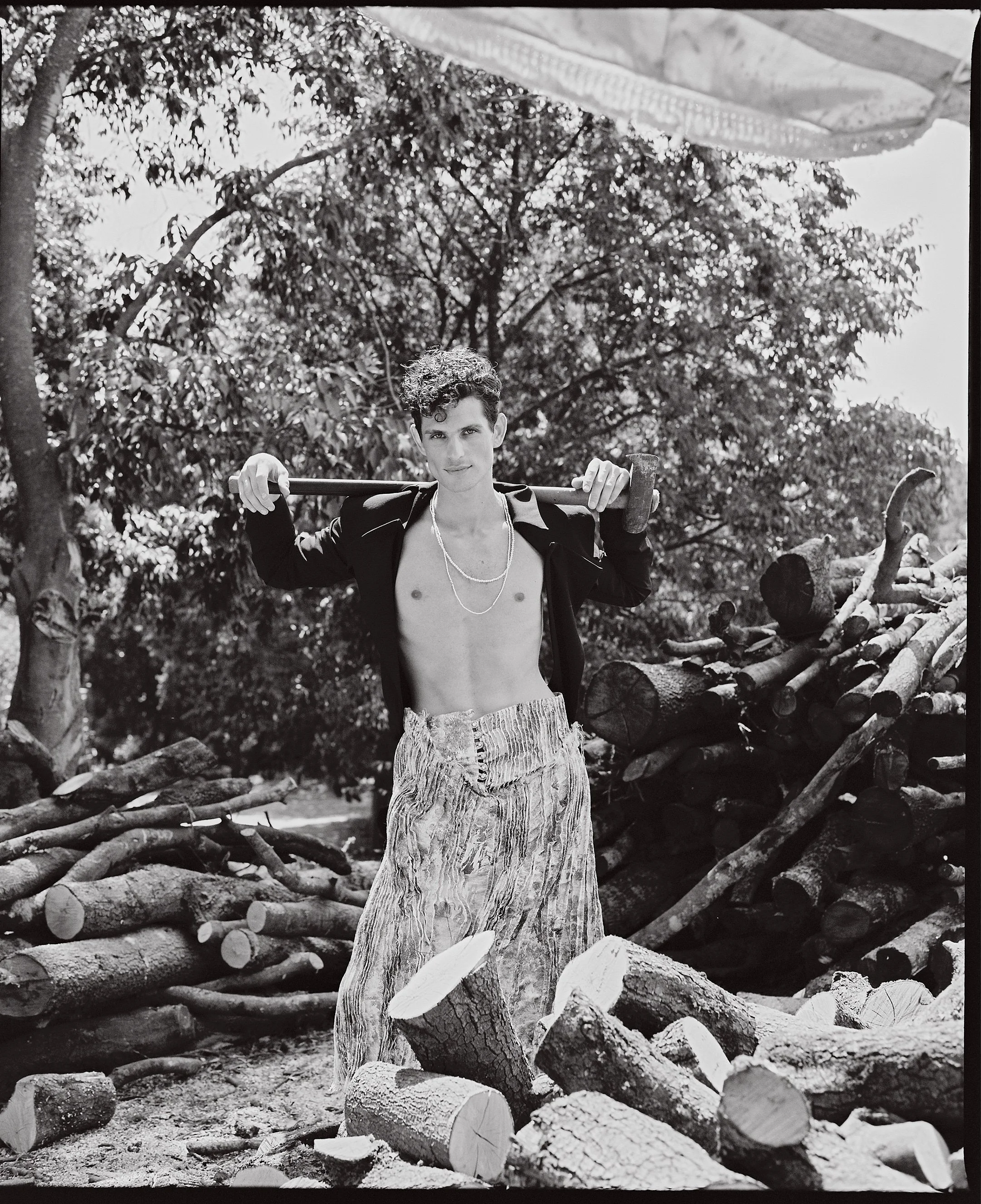 A young man with short curly hair, no shirt, wearing patterned pants, and a black jacket over his shoulders, posing outdoors with a large axe resting on his shoulders amidst stacked logs and trees.