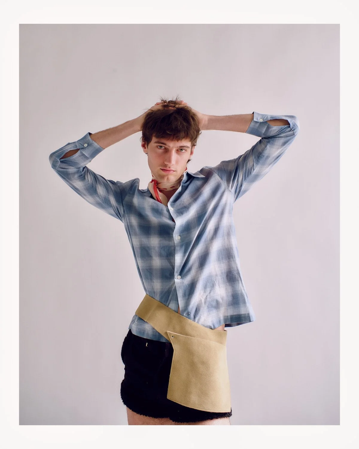 A young man with brown hair posing with hands behind his head, wearing a blue checkered shirt, a beige apron, and a black skirt, against a plain light background.