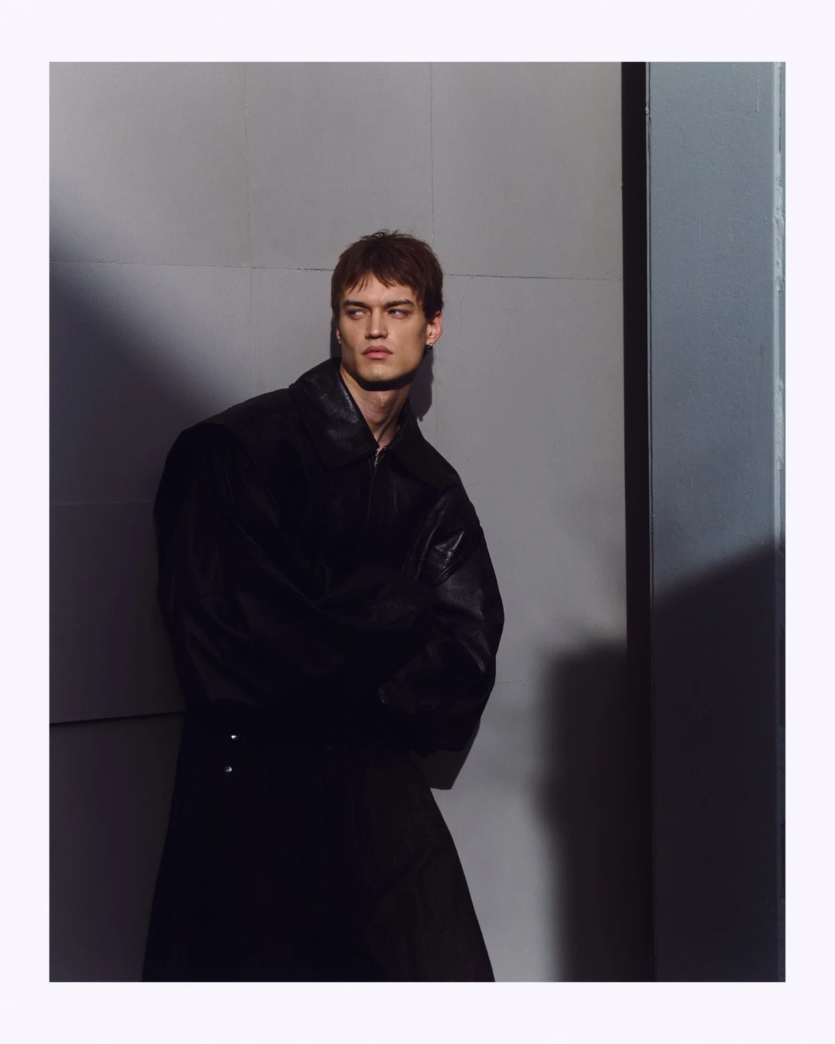 A young man with brown hair in a black leather jacket standing against a gray wall with shadowed lighting.