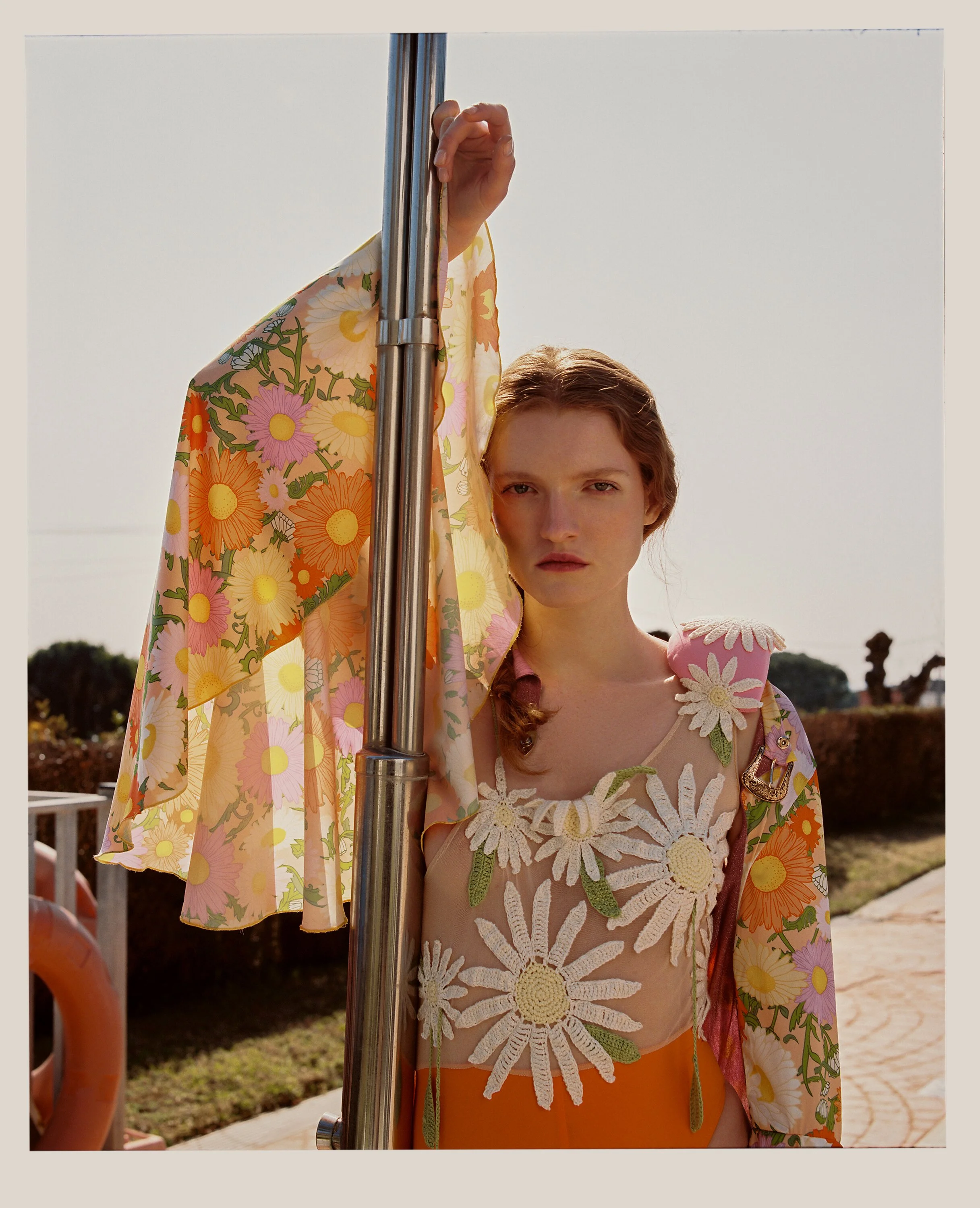 A young woman with red hair and fair skin standing outdoors, holding onto a metal pole with her left hand. She is wearing a sheer top decorated with large white crochet daisies with yellow centers and green leaves, and a brightly colored floral jacke