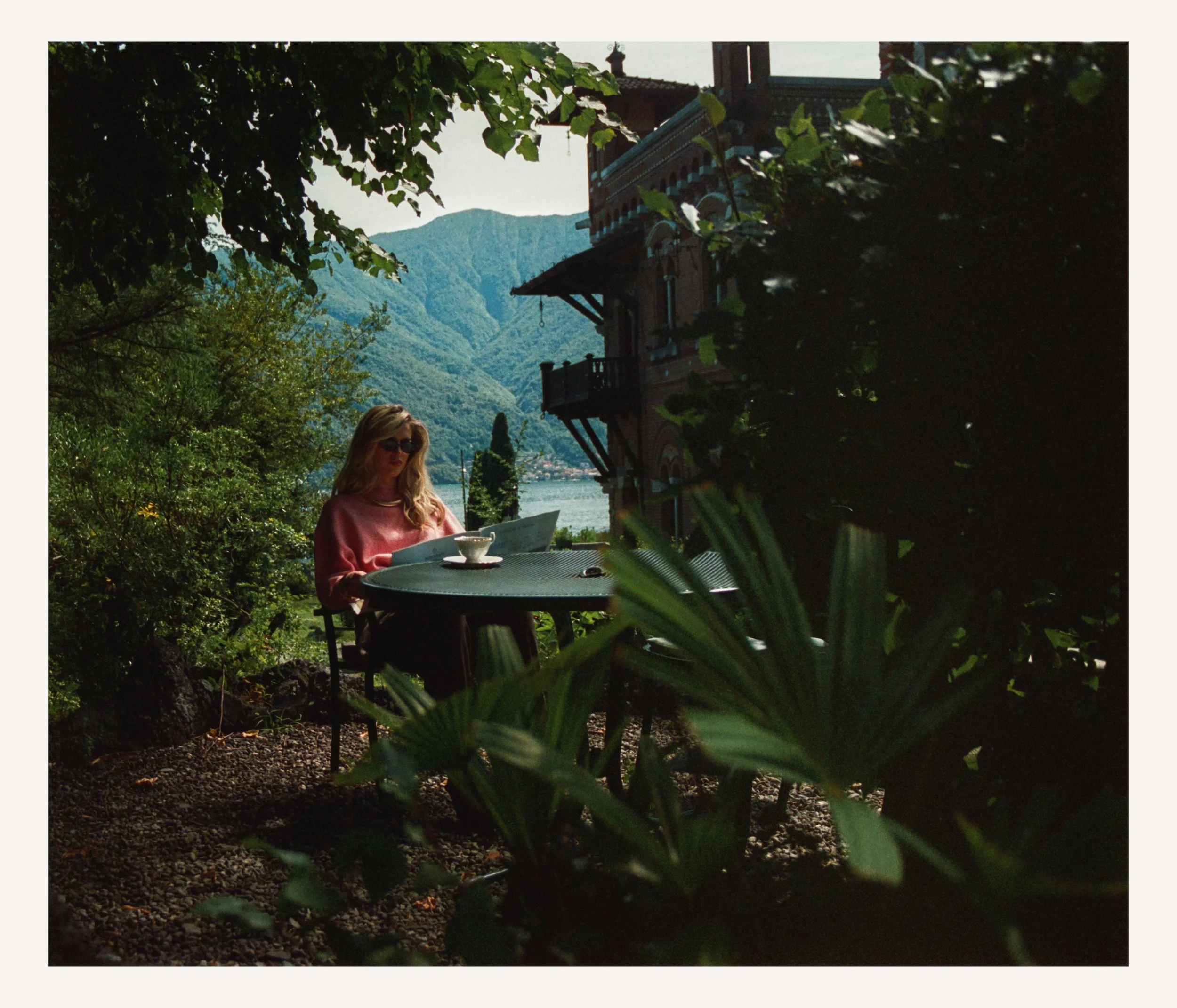 A woman sitting at a round table outdoors in a garden, with a lake and mountains in the background, surrounded by lush green foliage.