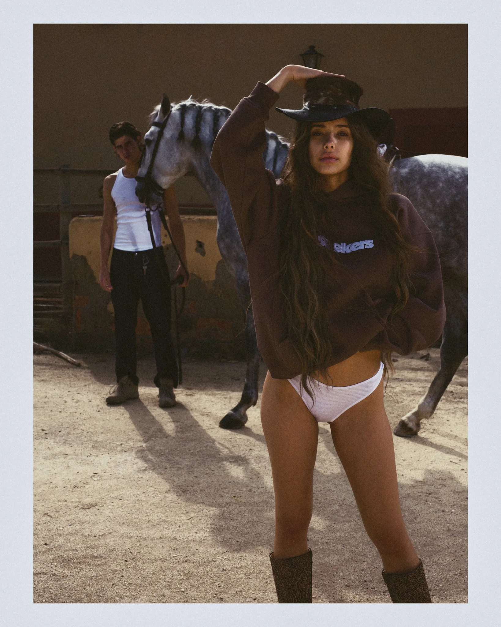 Young woman in a cowboy hat, hoodie, and white underwear standing outdoors near a horse with a man in the background.