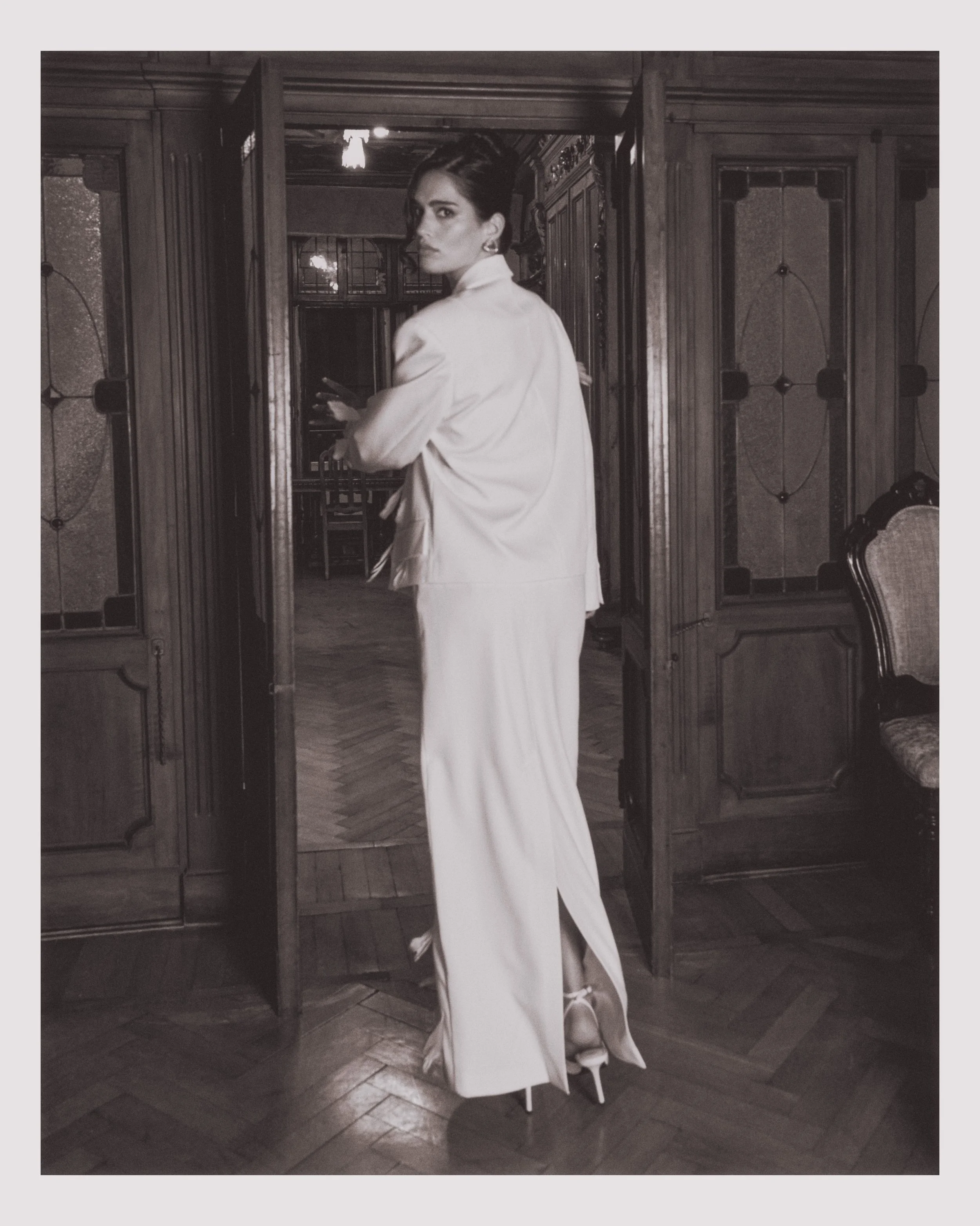 A woman in a white suit and high heels standing on stilts, looking over her shoulder in a wood-paneled room.
