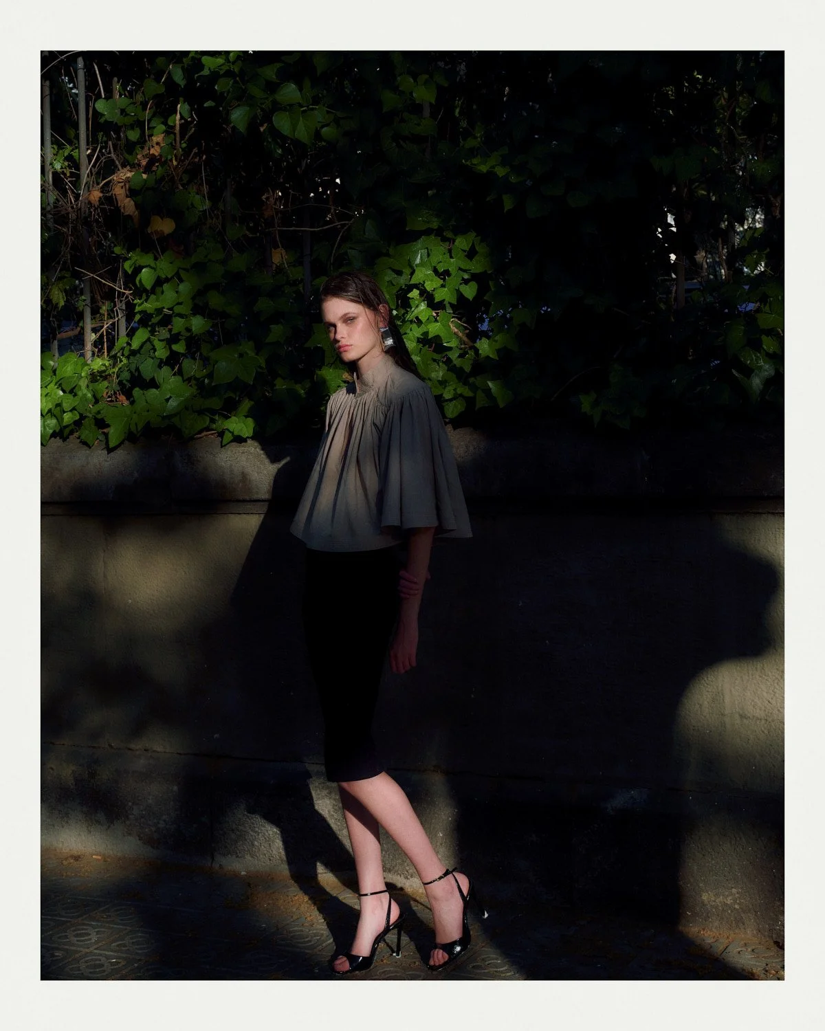 A woman in a beige top and black skirt standing outdoors against a background of green foliage and a concrete wall, with sunlight creating shadows.
