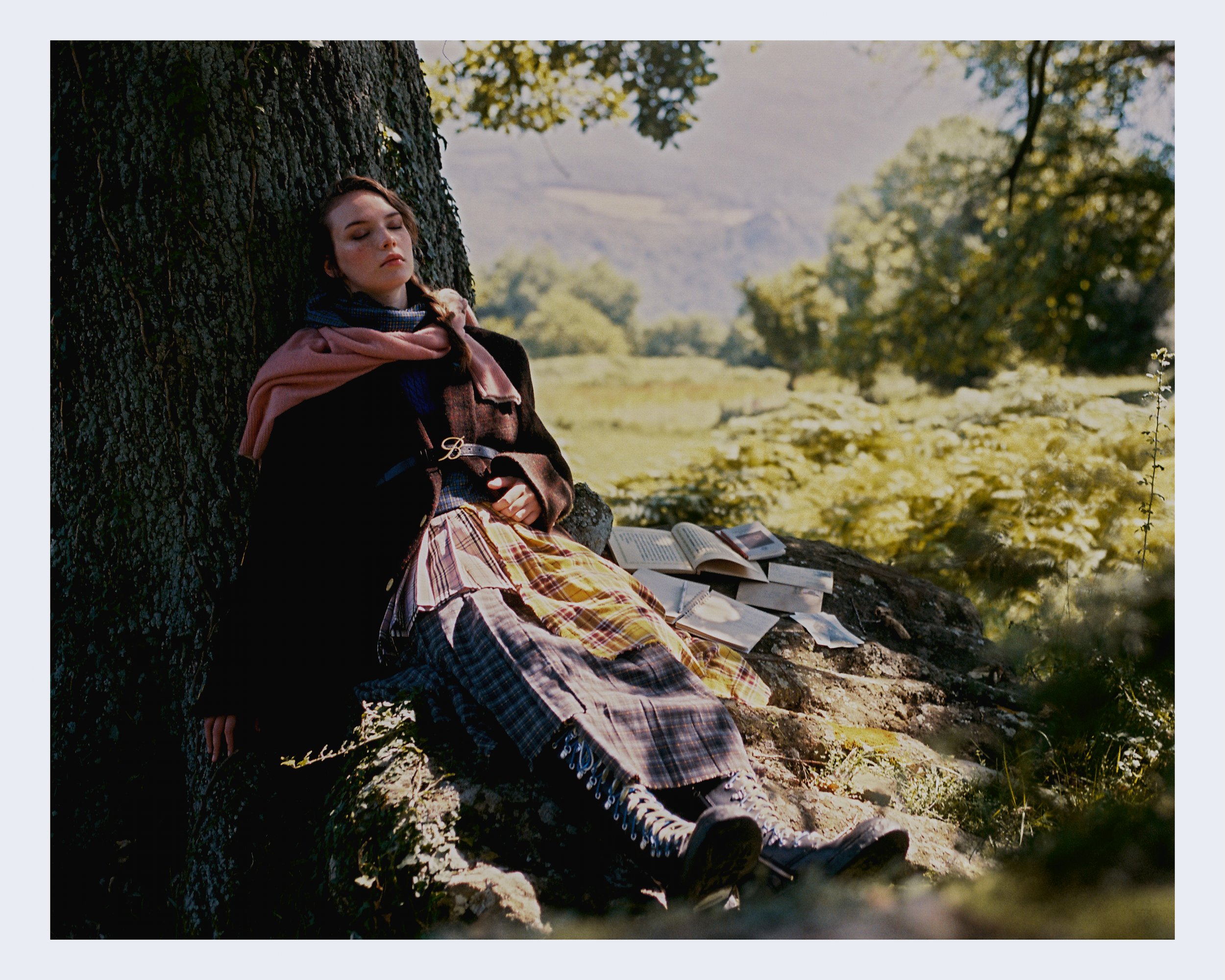 Young woman with long brown hair, wearing a plaid skirt, black jacket, and a pink and blue scarf, sits against a large tree in a sunlit forest, with open books on the ground beside her, appearing to be resting or daydreaming.